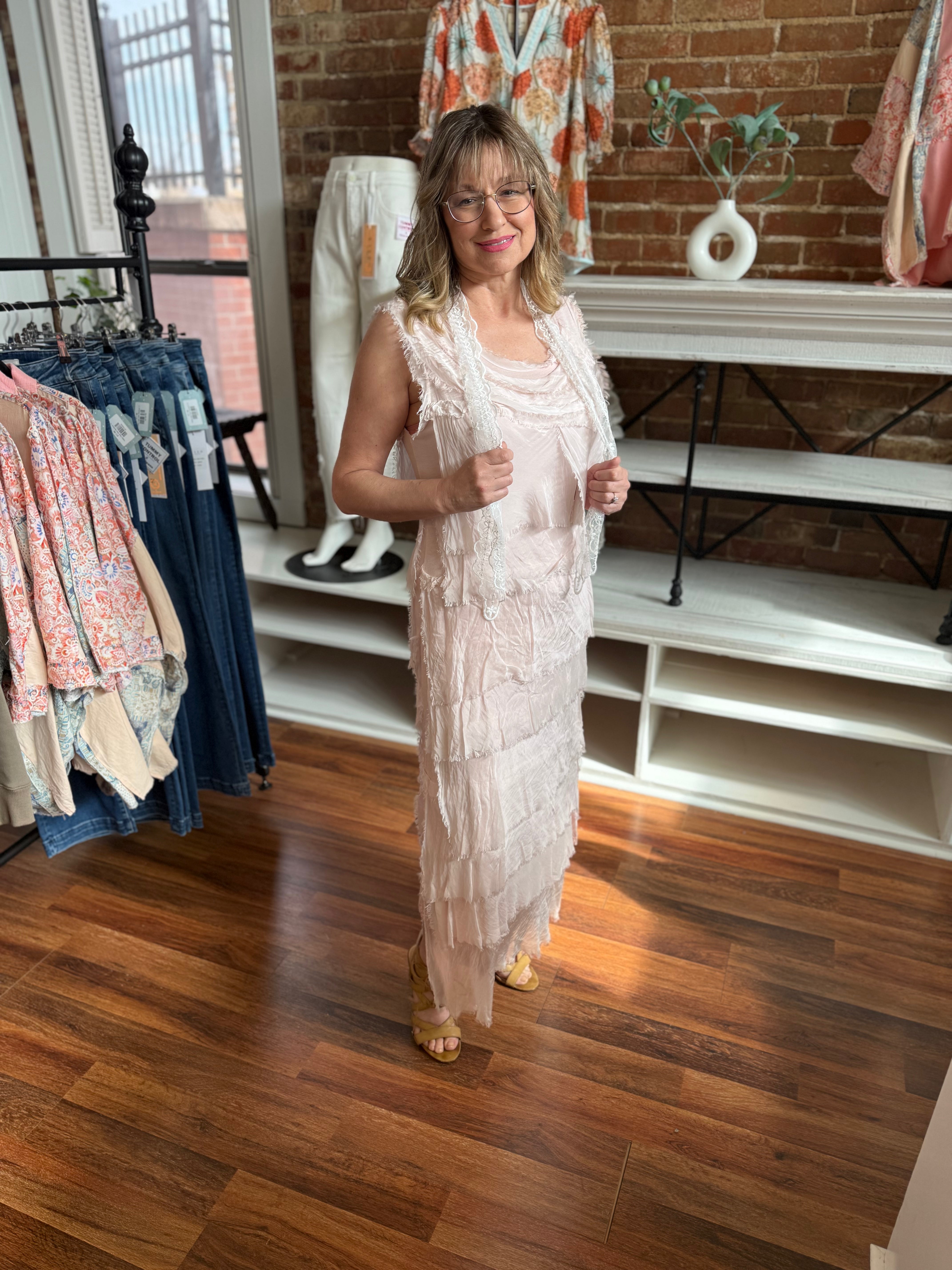 Woman in a light pink dress standing in a clothing store with racks of clothes and a brick wall in the background.