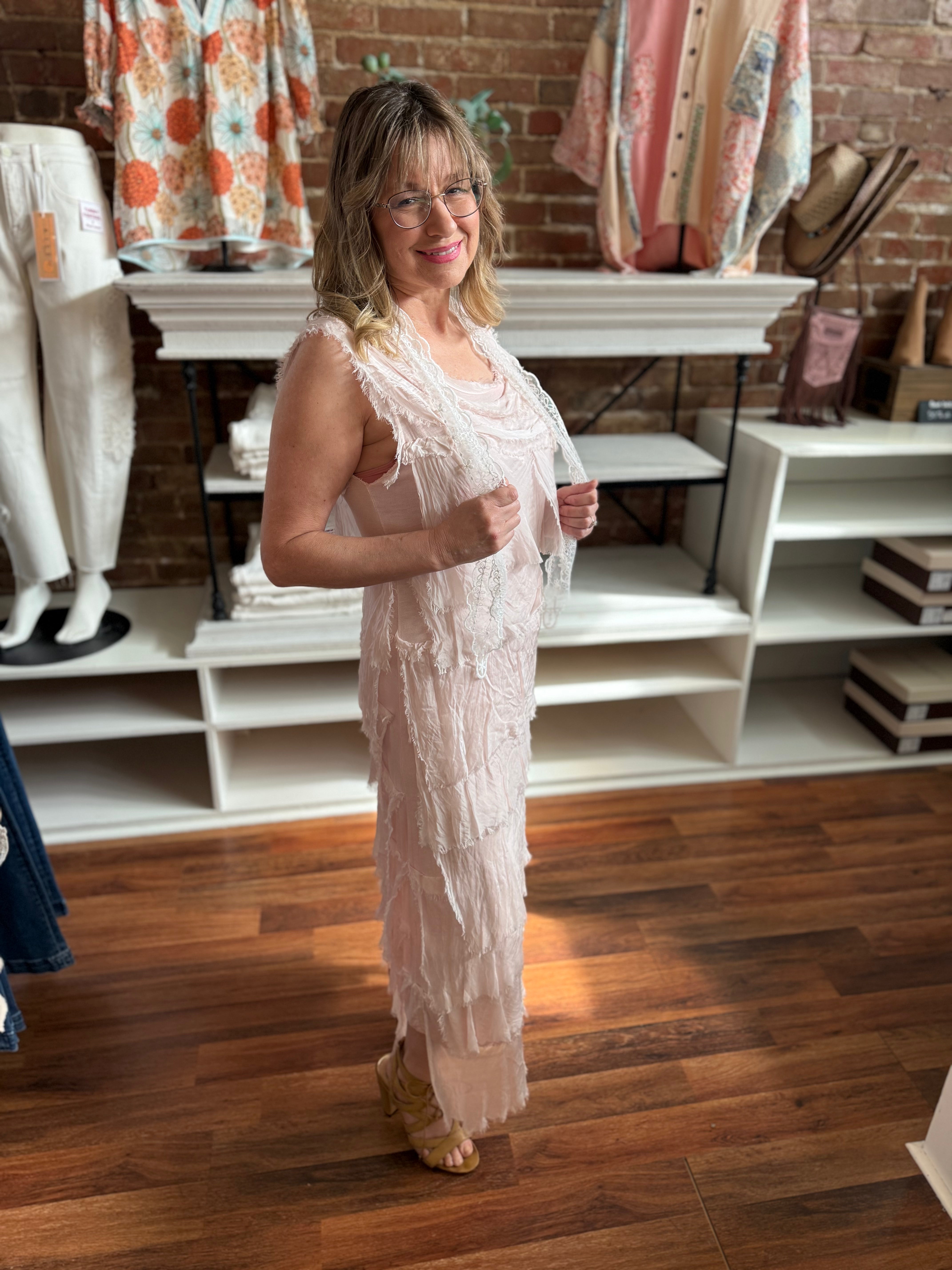 Woman in a light pink dress standing in a store with clothing racks and shelves in the background.