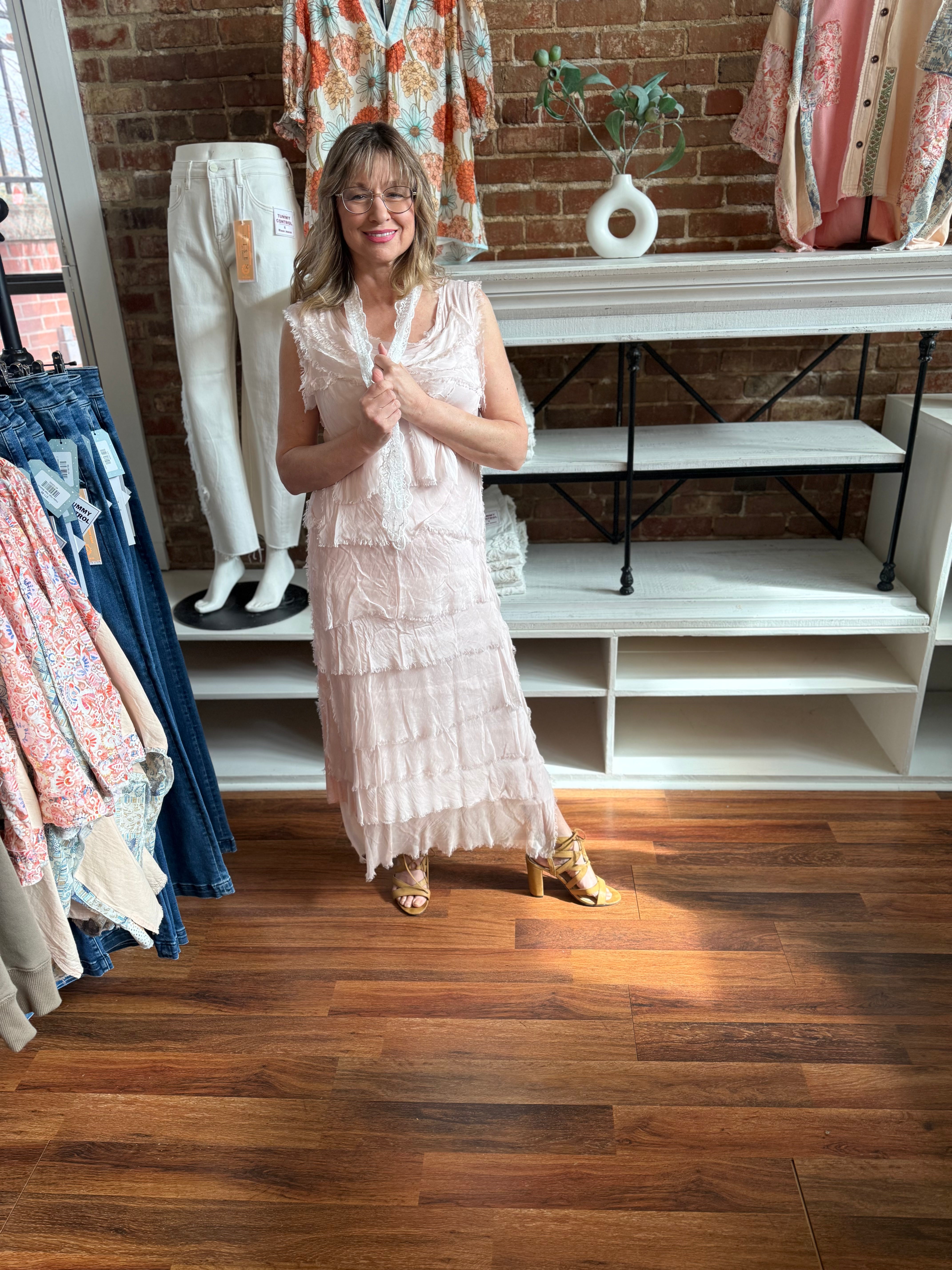 Woman in a light pink dress standing in a clothing store with racks of clothes and a brick wall background.