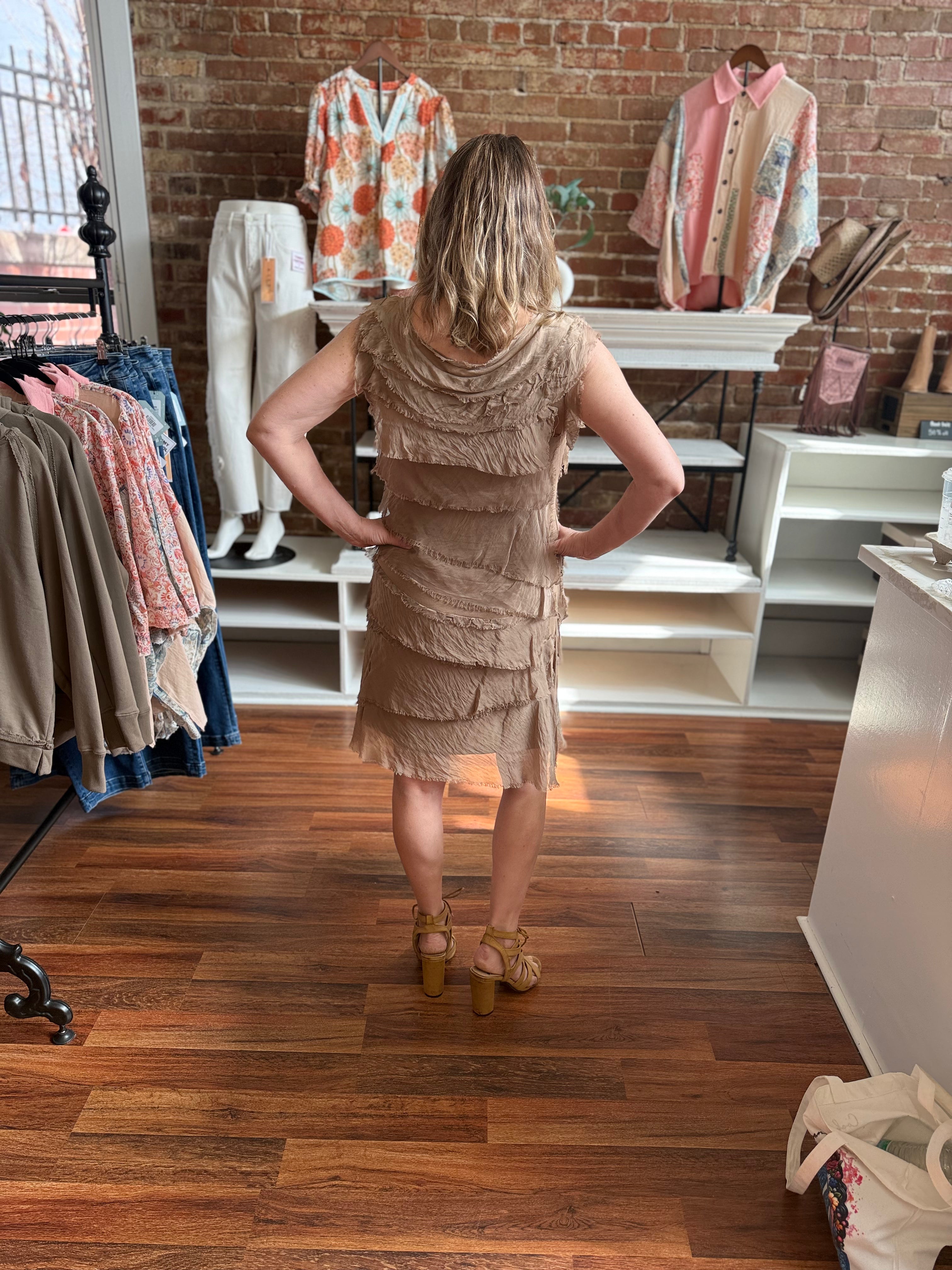 Woman in a beige dress standing in a clothing store with racks of clothes and a brick wall.