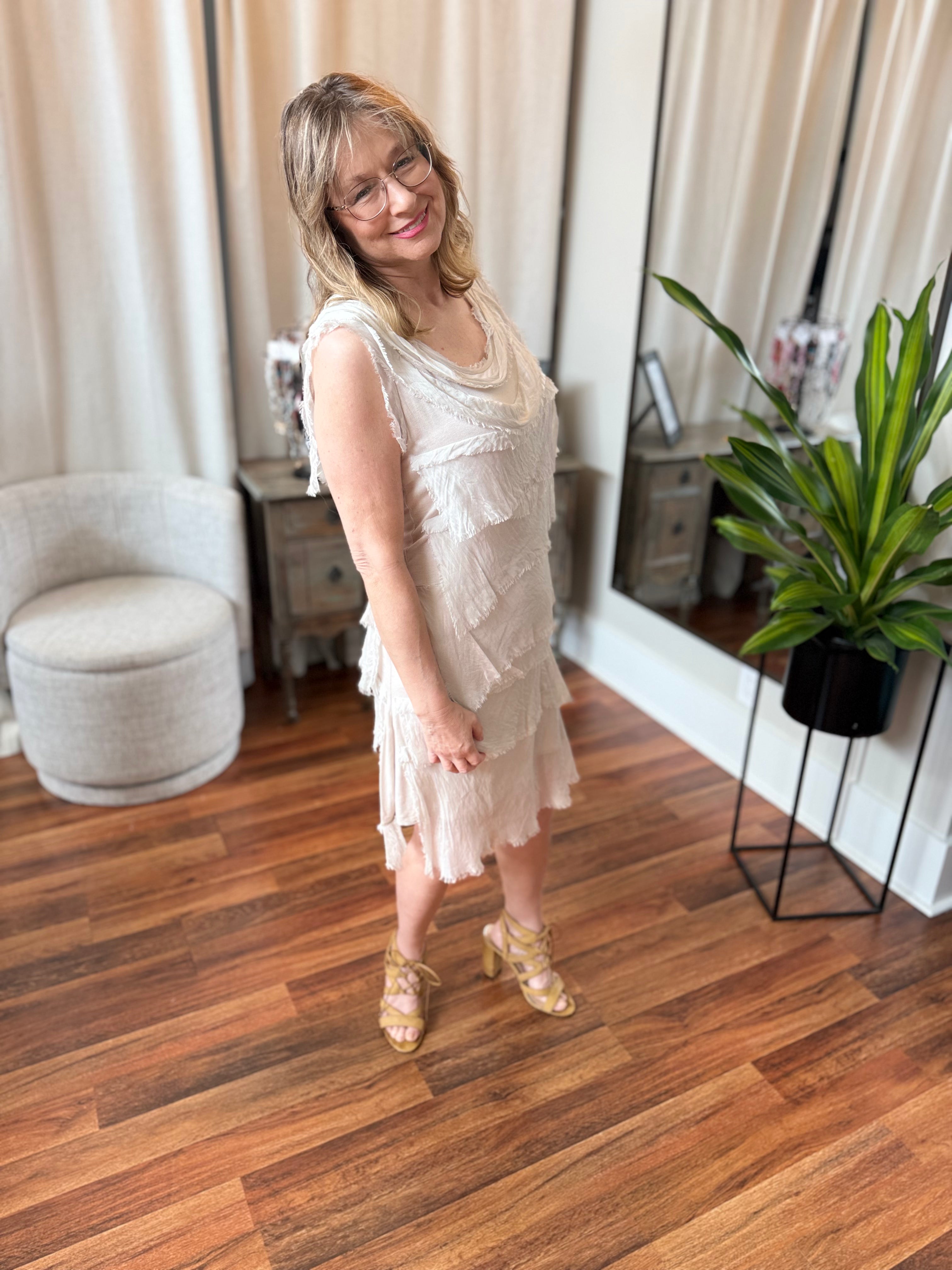 Woman in a white dress standing in a room with wooden flooring and a plant.