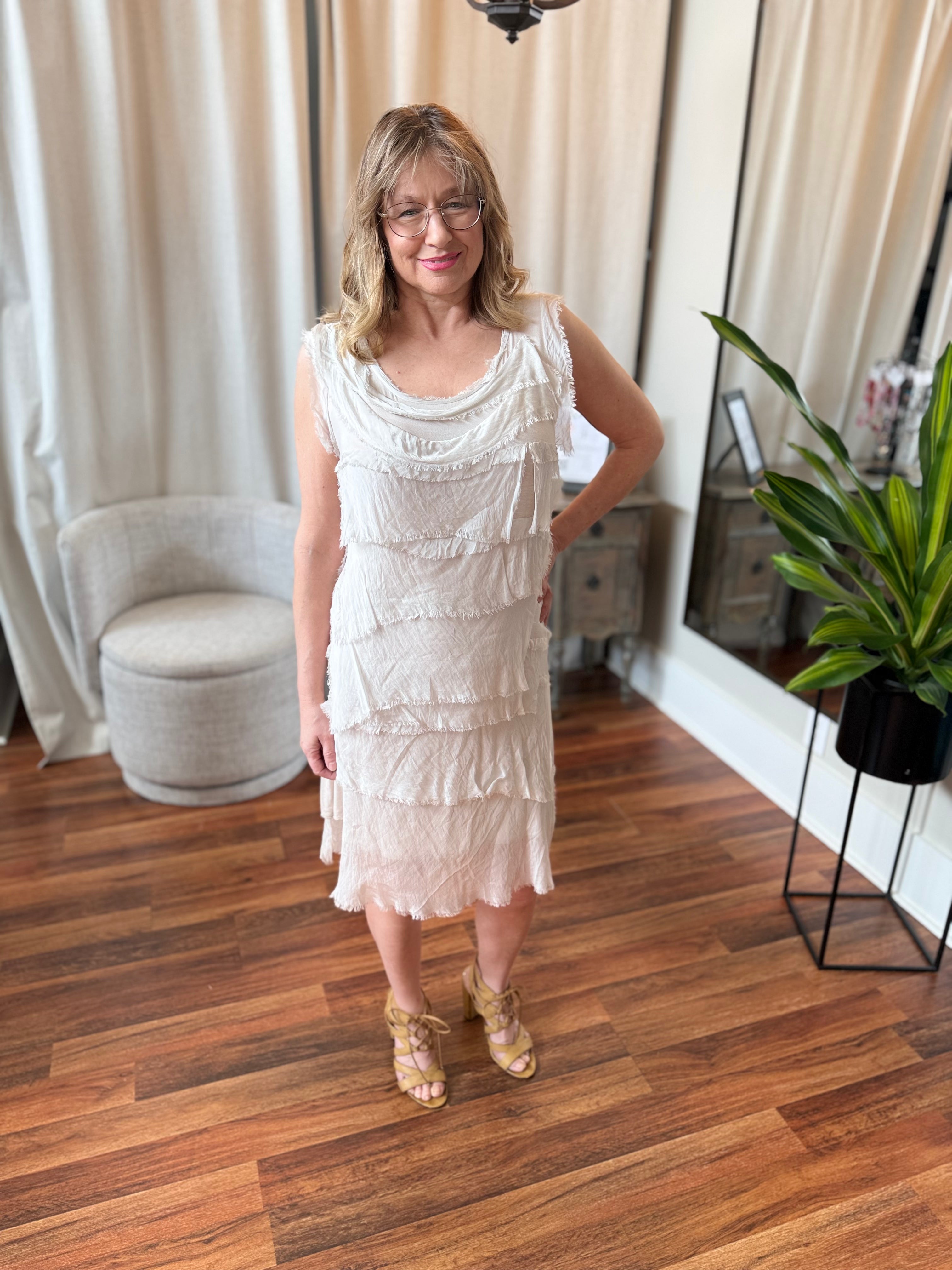 Woman in a white dress standing in a room with wooden flooring and curtains.