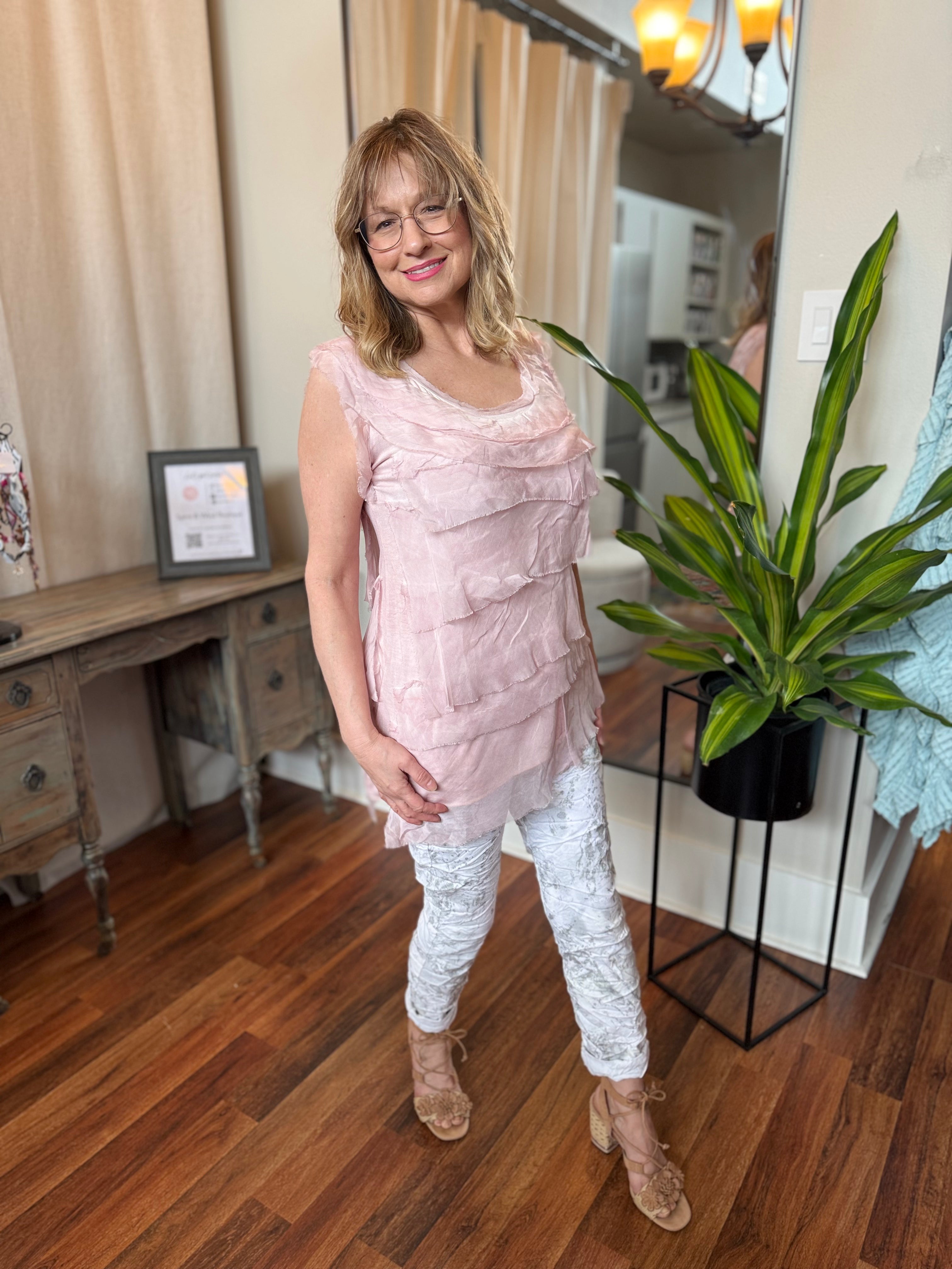 Woman in a pink top and white pants standing in a room with wooden floor and plant.