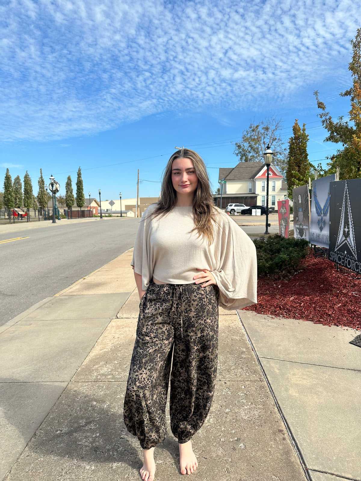 A woman stands on a sidewalk outdoors wearing a cream-colored draped top and dark patterned harem pants under a blue sky.