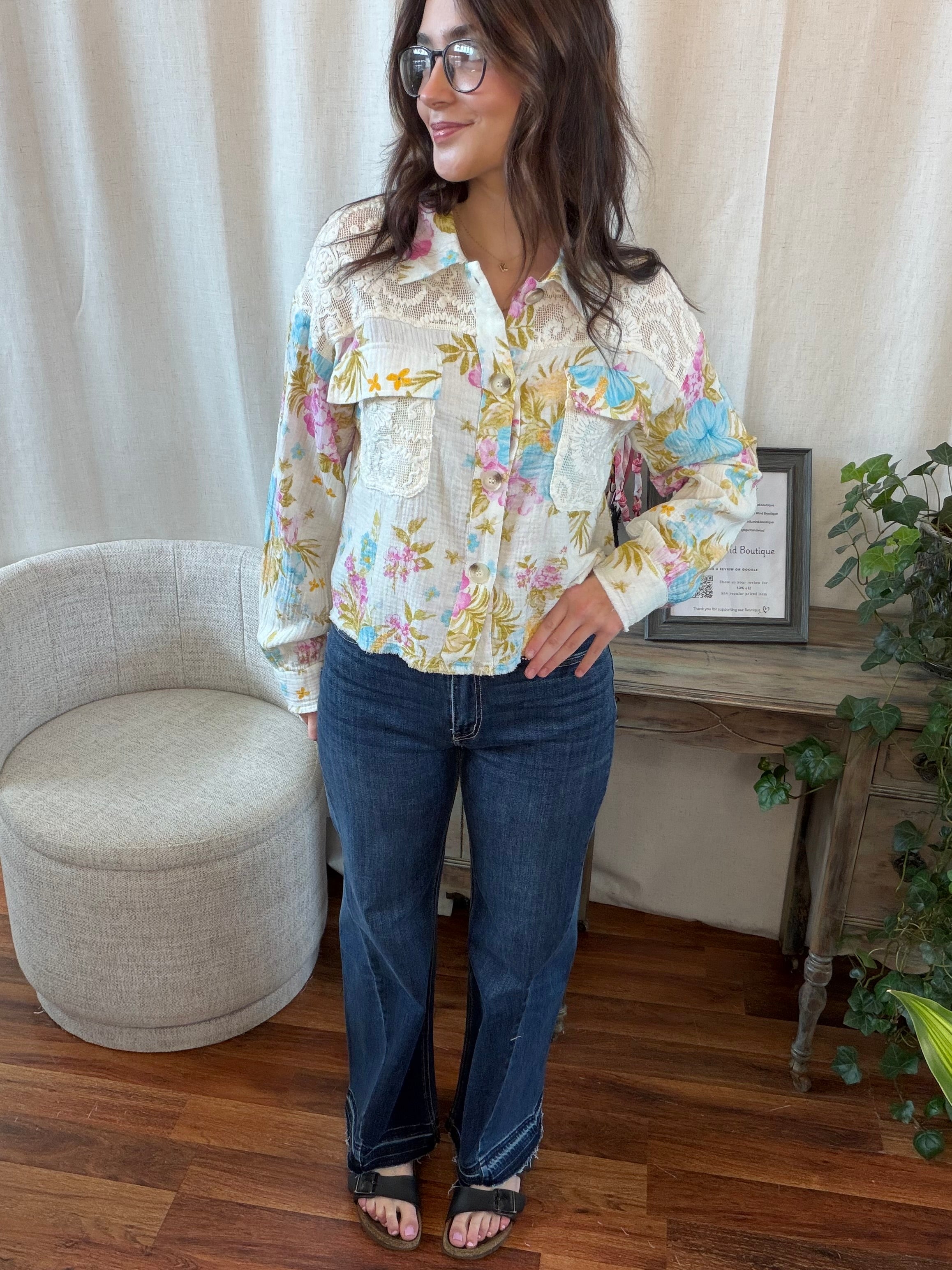 Woman wearing a floral blouse and blue jeans standing in a room with a chair and plant.