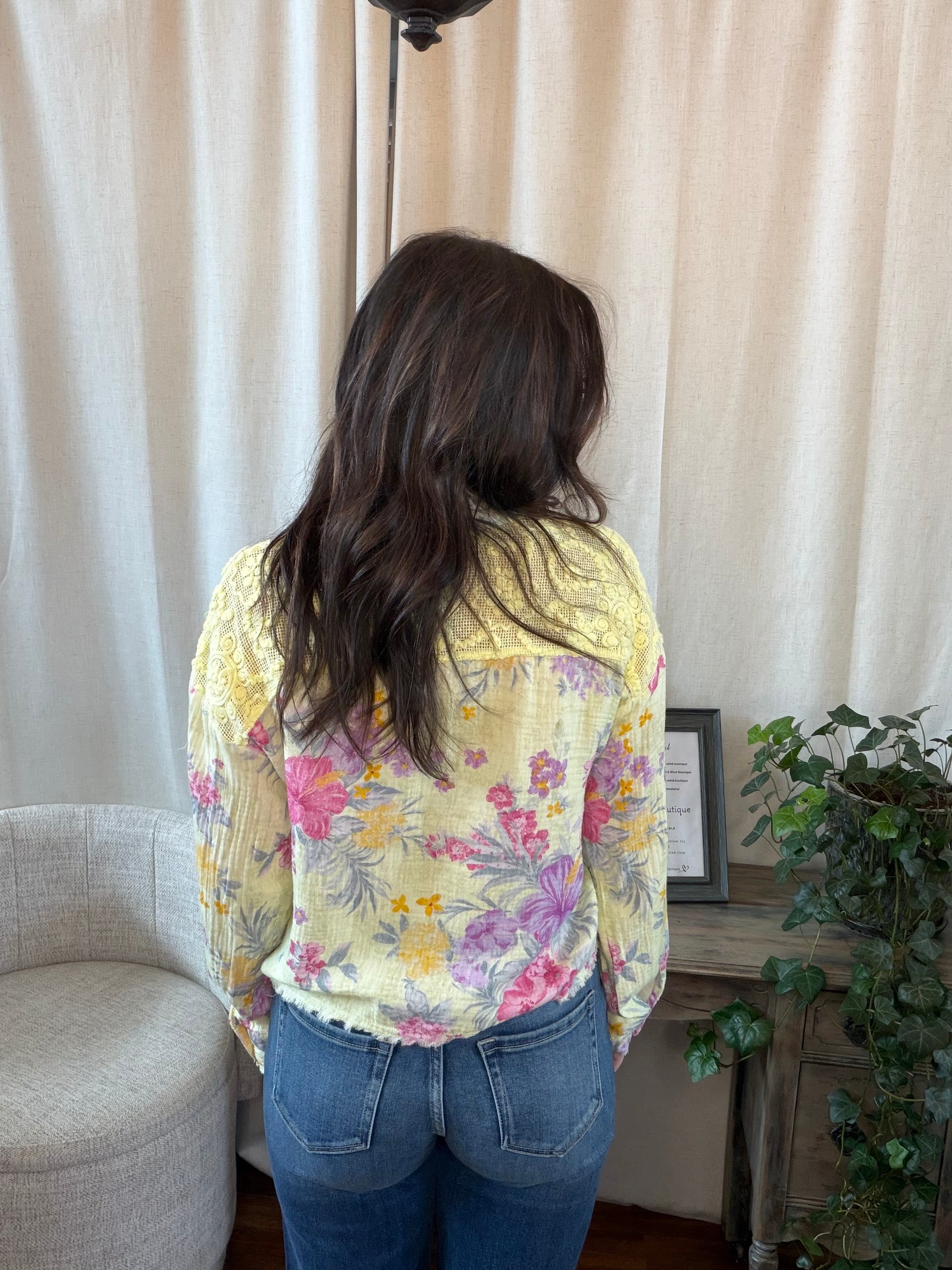 Person wearing a floral shirt and yellow jacket in a room with curtains and a plant.