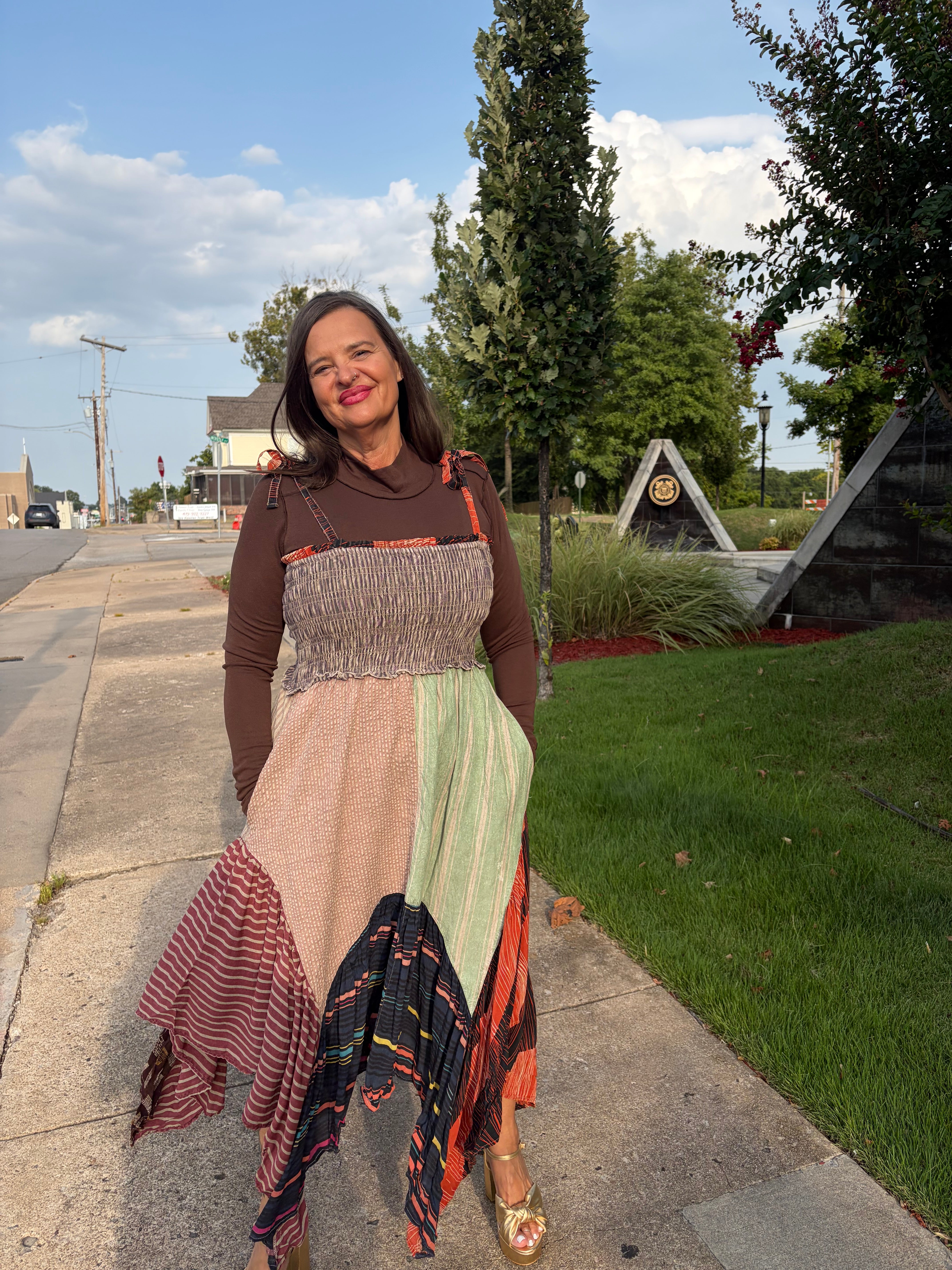 A woman wearing a long, patchwork-style maxi dress stands on a sidewalk outdoors. The dress has multiple panels in earthy tones including brown, beige, sage green, orange, and black with red accents.