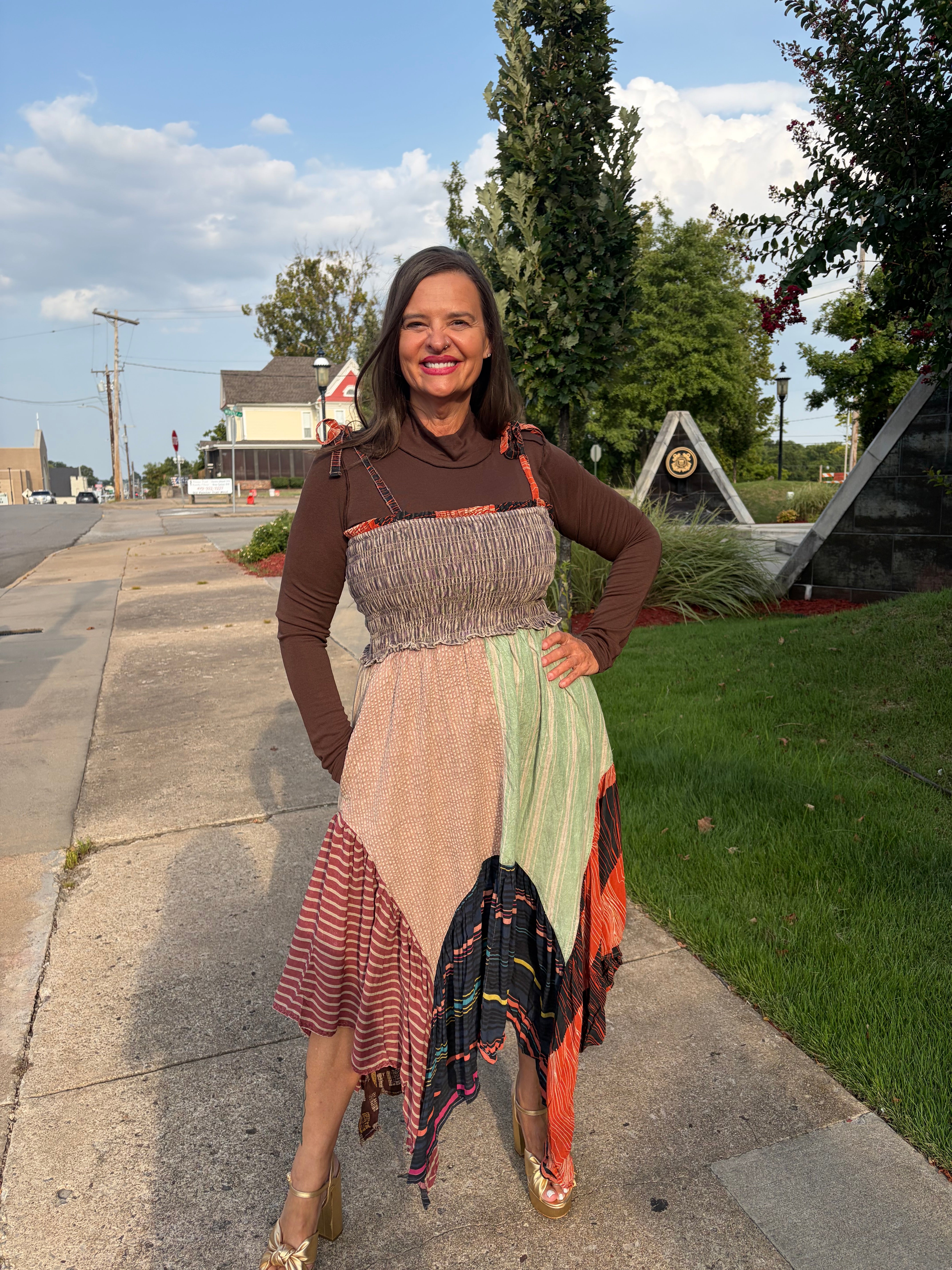 A woman stands on a sidewalk outdoors wearing a patchwork midi dress with a brown long-sleeve top underneath. The dress features multiple fabric panels in beige, light green, orange, red, and black, with a smocked bodice.