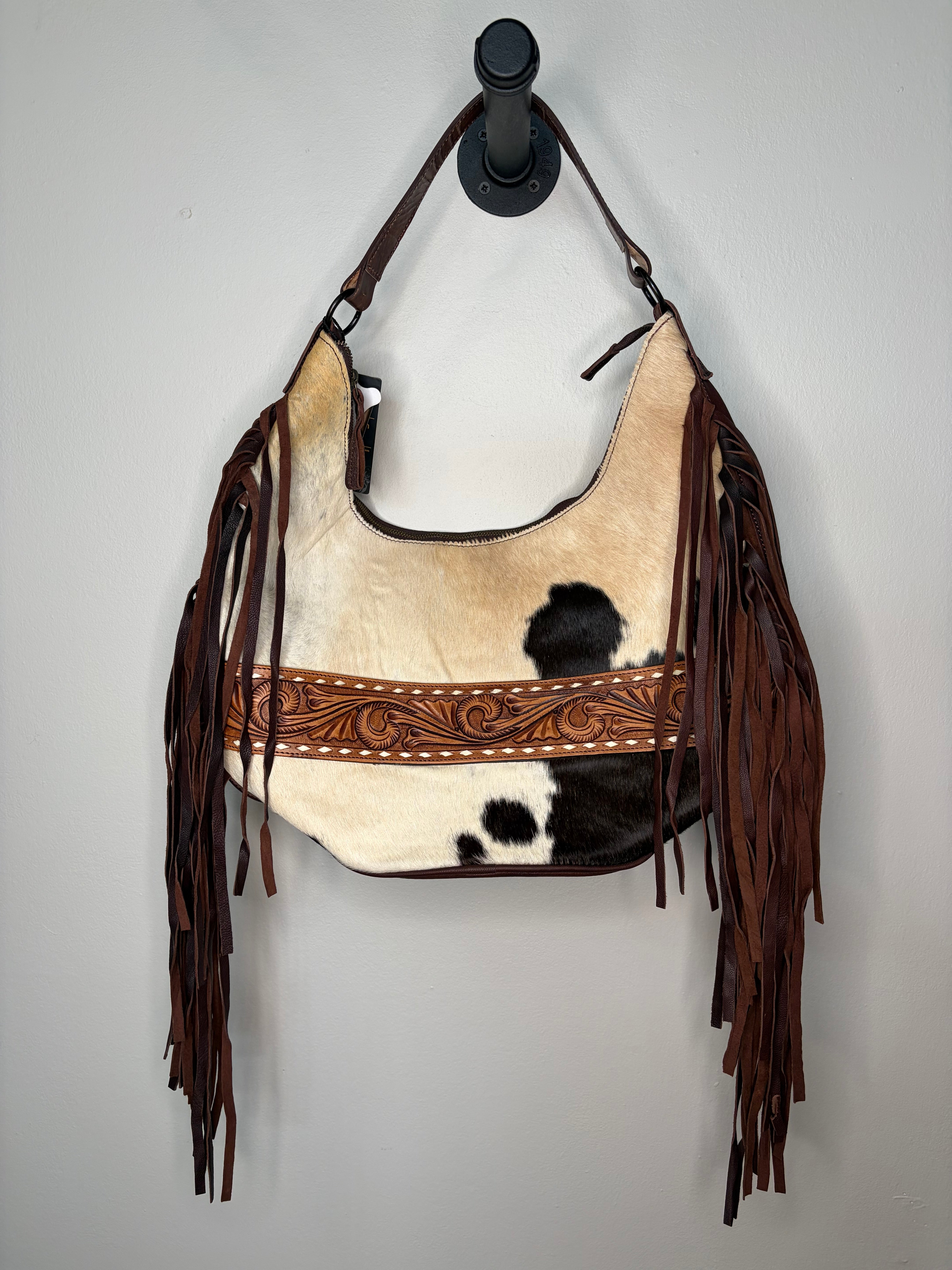 A western-style shoulder bag made from cowhide and brown leather fringe, hanging from a black wall hook against a light gray wall. The bag features a natural black-and-white cowhide body, a tooled brown leather band across the front, and long dark brown leather fringe on the sides.