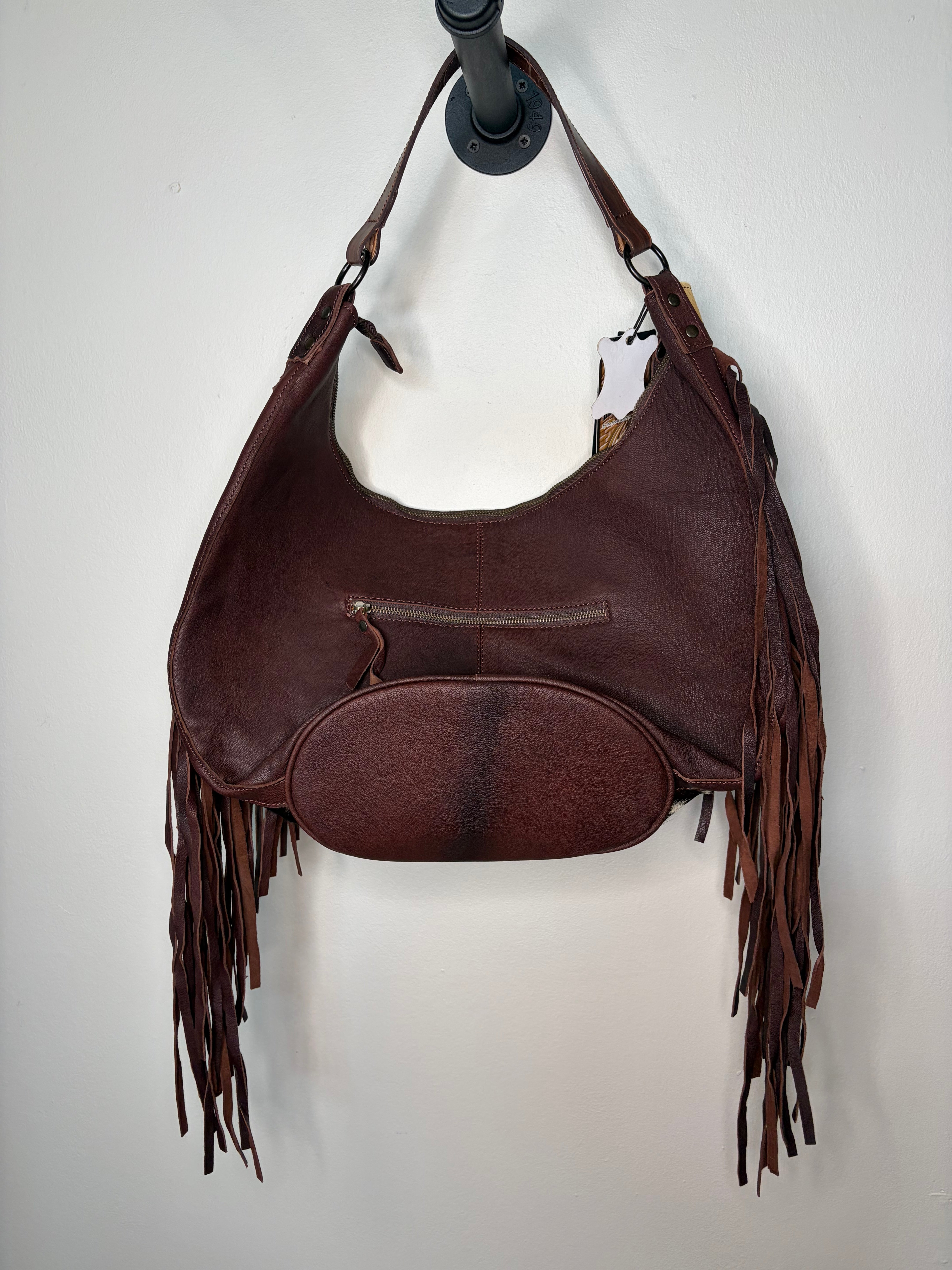 A dark brown leather shoulder bag with fringe details hanging from the sides, shown hanging from a black wall hook against a plain off-white wall.