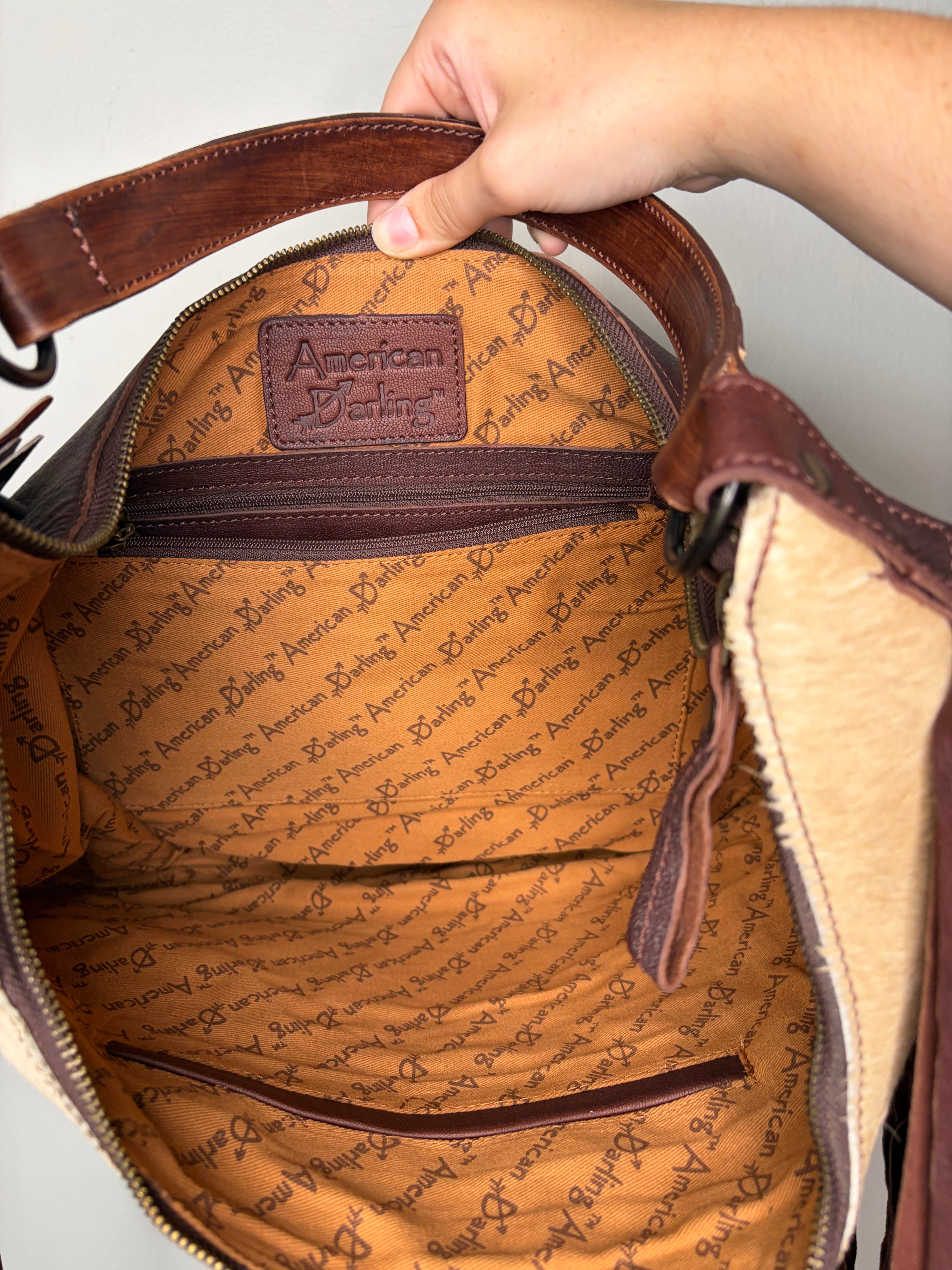An open brown leather handbag shown from the inside, with a hand holding the handle. The interior is lined with tan patterned fabric and features a brown leather patch with the embossed brand name 'American Eagle'.