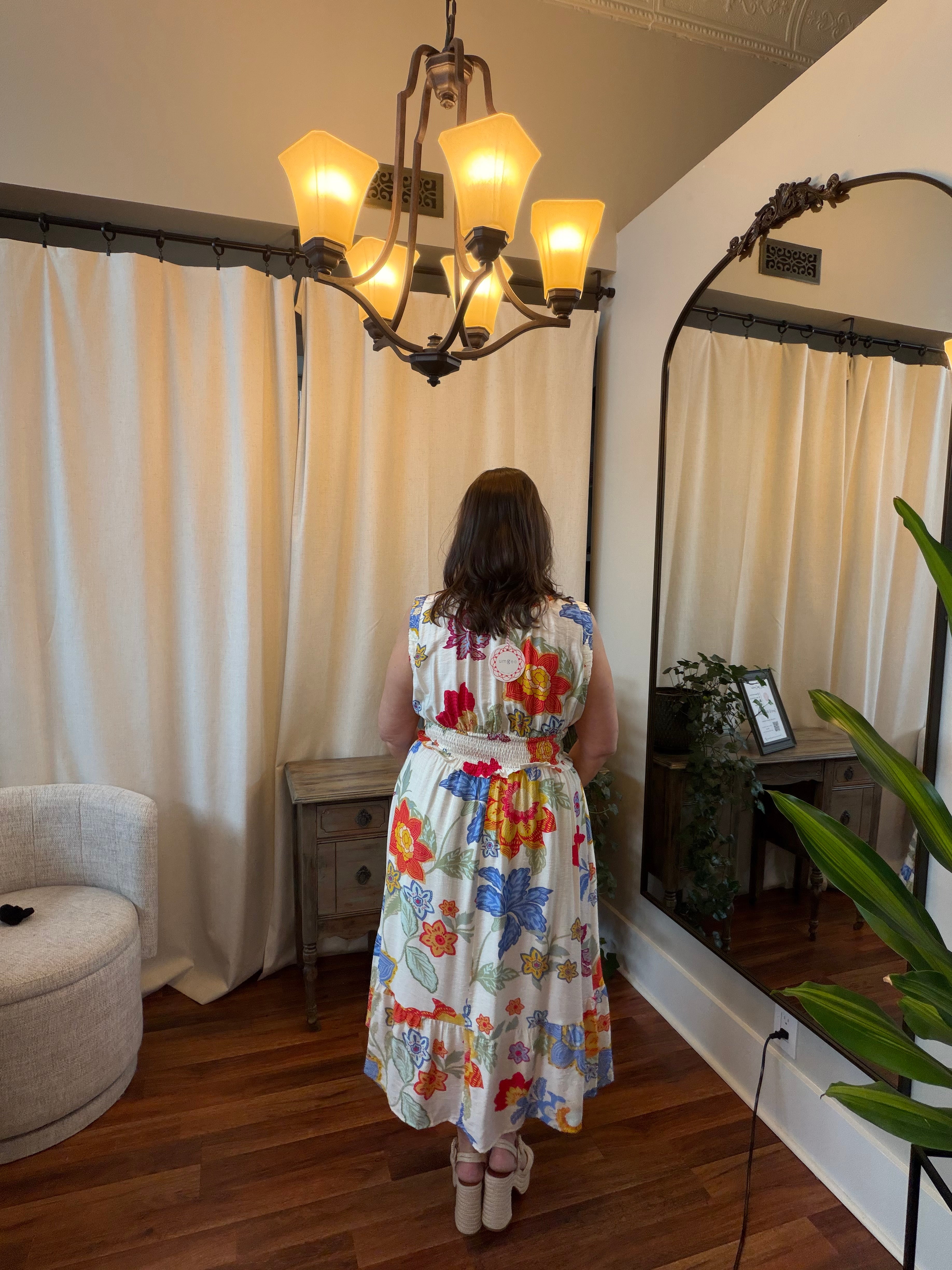 Woman in a floral dress standing in a room with a chandelier and large mirror.