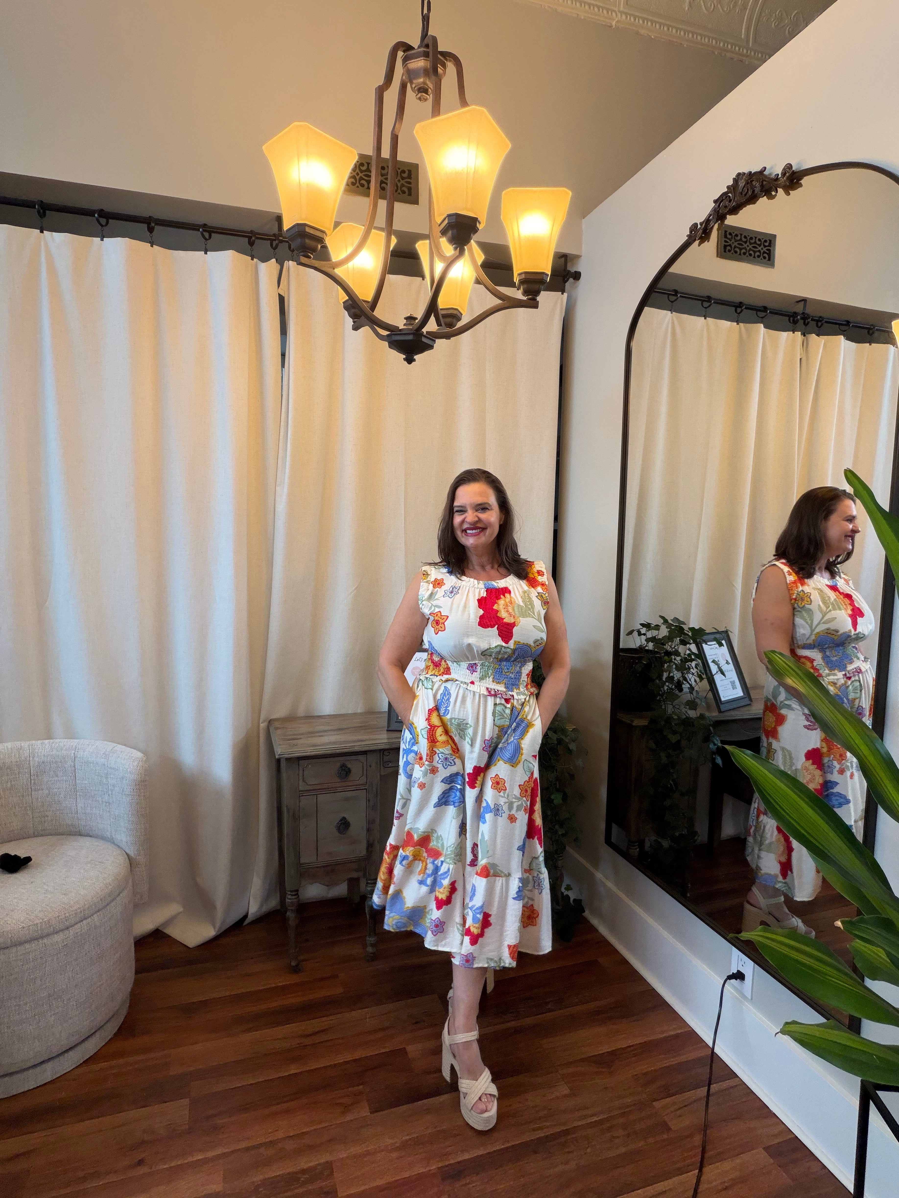 Woman in a floral dress standing in a room with a chandelier and large mirror.