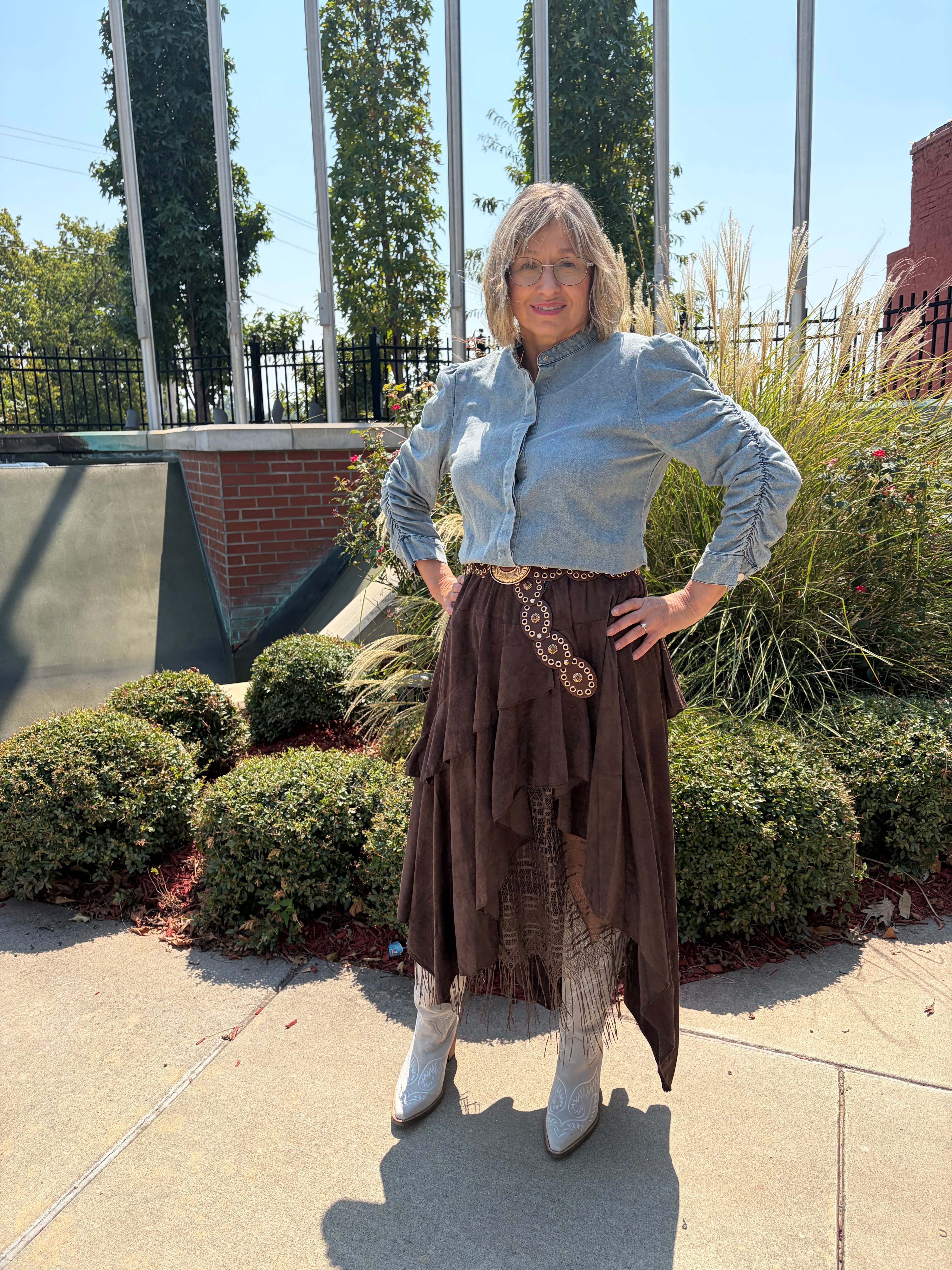  — Boho Layered Lace Skirt in dark chocolate brown with asymmetrical layered hem and lace fringe, styled with light blue denim top and white cowboy boots, front view outdoors
