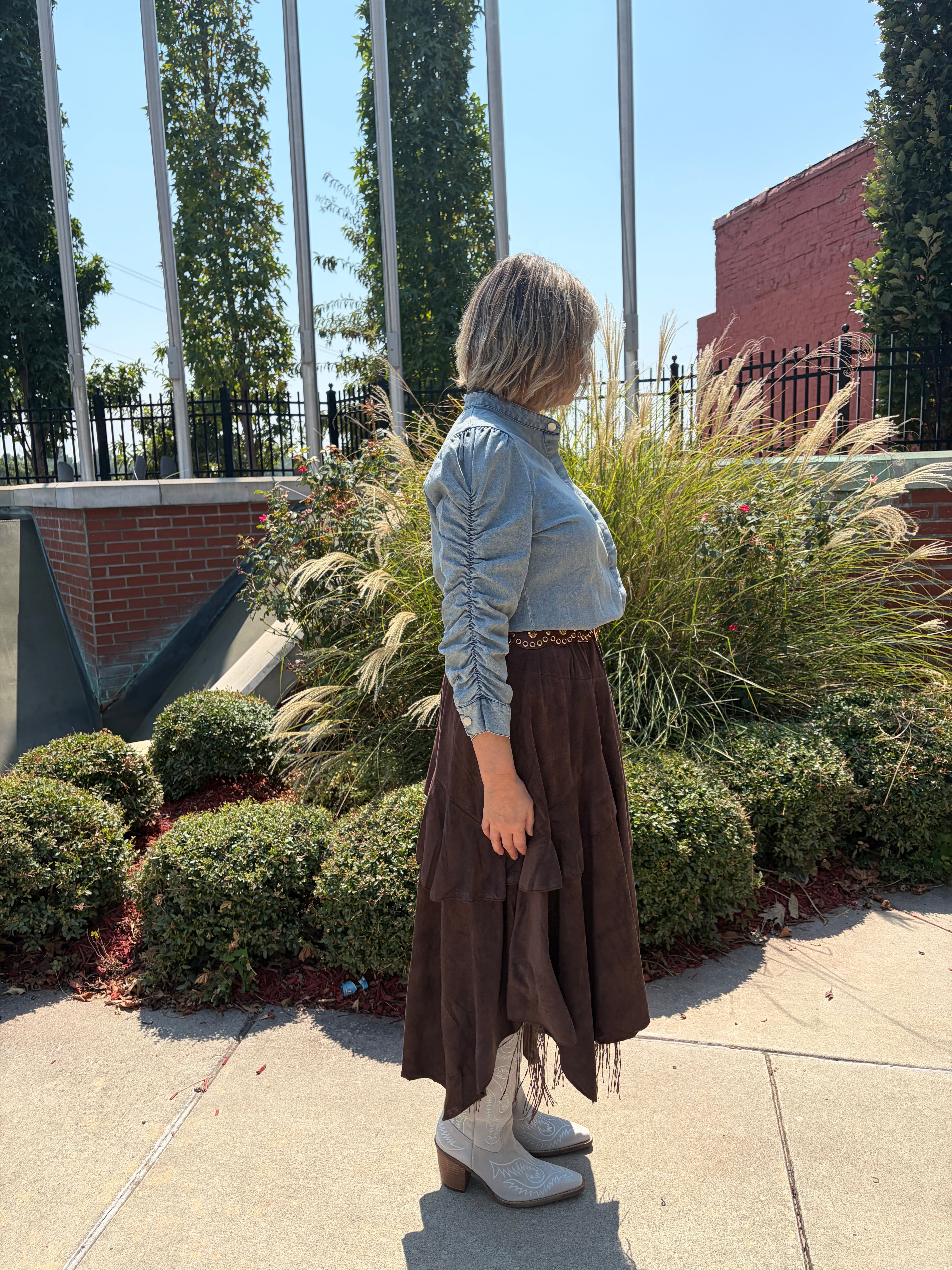 A woman stands outdoors on a sidewalk wearing a light blue long-sleeve ruched shirt and a brown tiered midi skirt, paired with white heeled boots. The background features green landscaping, tall flagpoles, and a brick building under a clear blue sky.