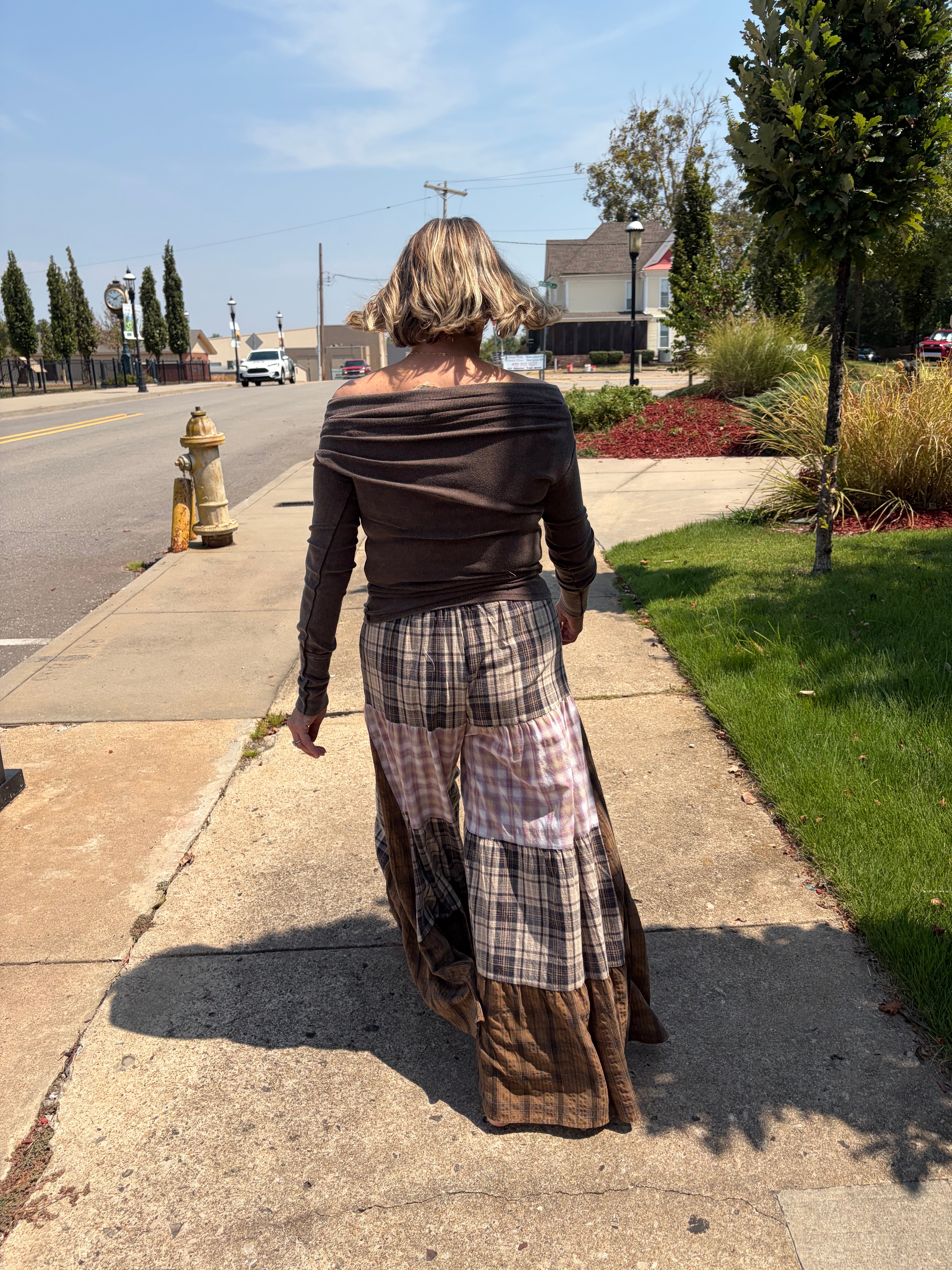 Patchwork Plaid Maxi Pants in tiered brown and neutral plaid panels, back view walking on sidewalk, styled with dark brown off-shoulder long sleeve top