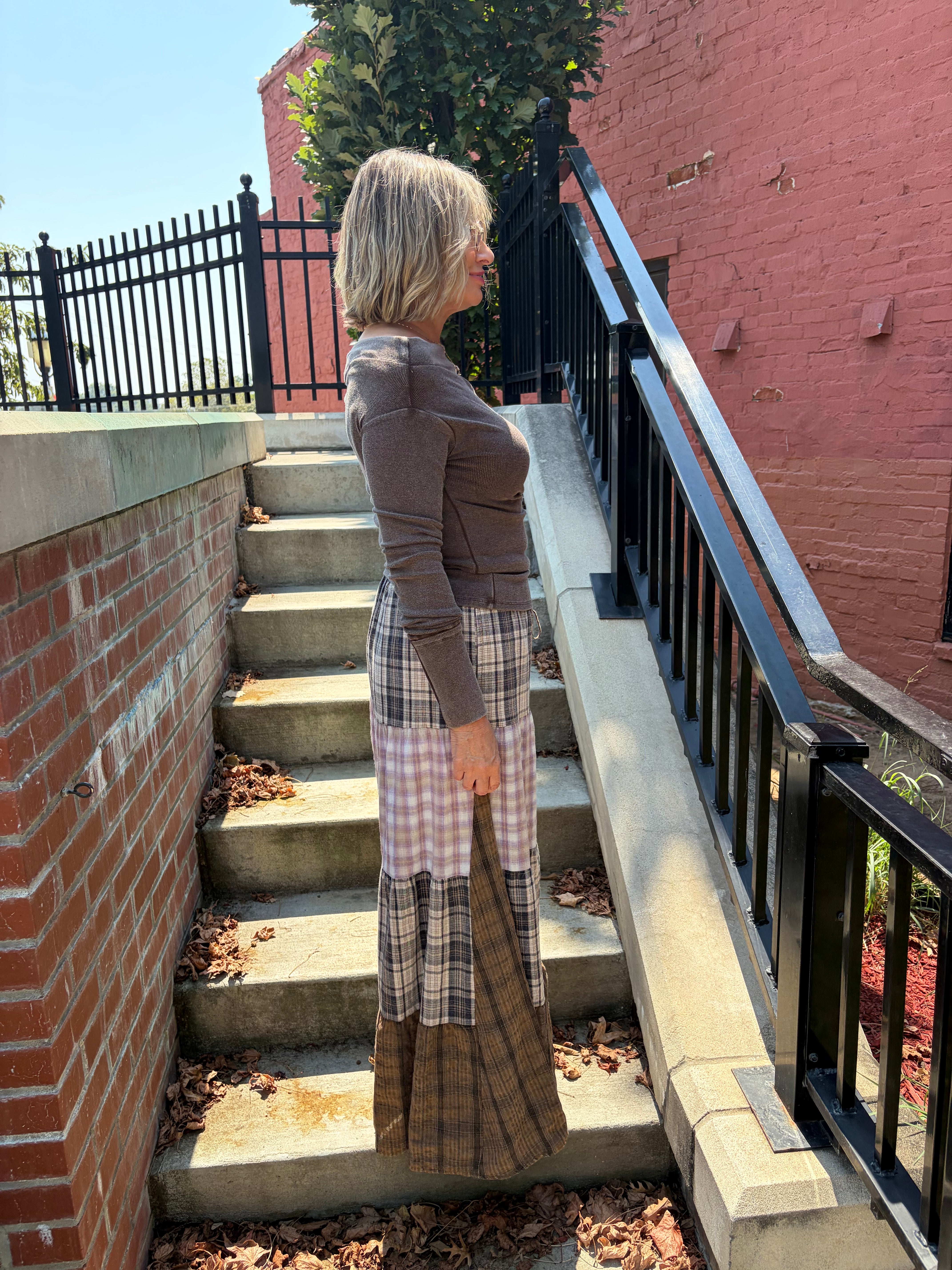 Patchwork Plaid Maxi Pants in tiered brown plaid patchwork, side profile view on outdoor staircase, styled with brown long sleeve top
