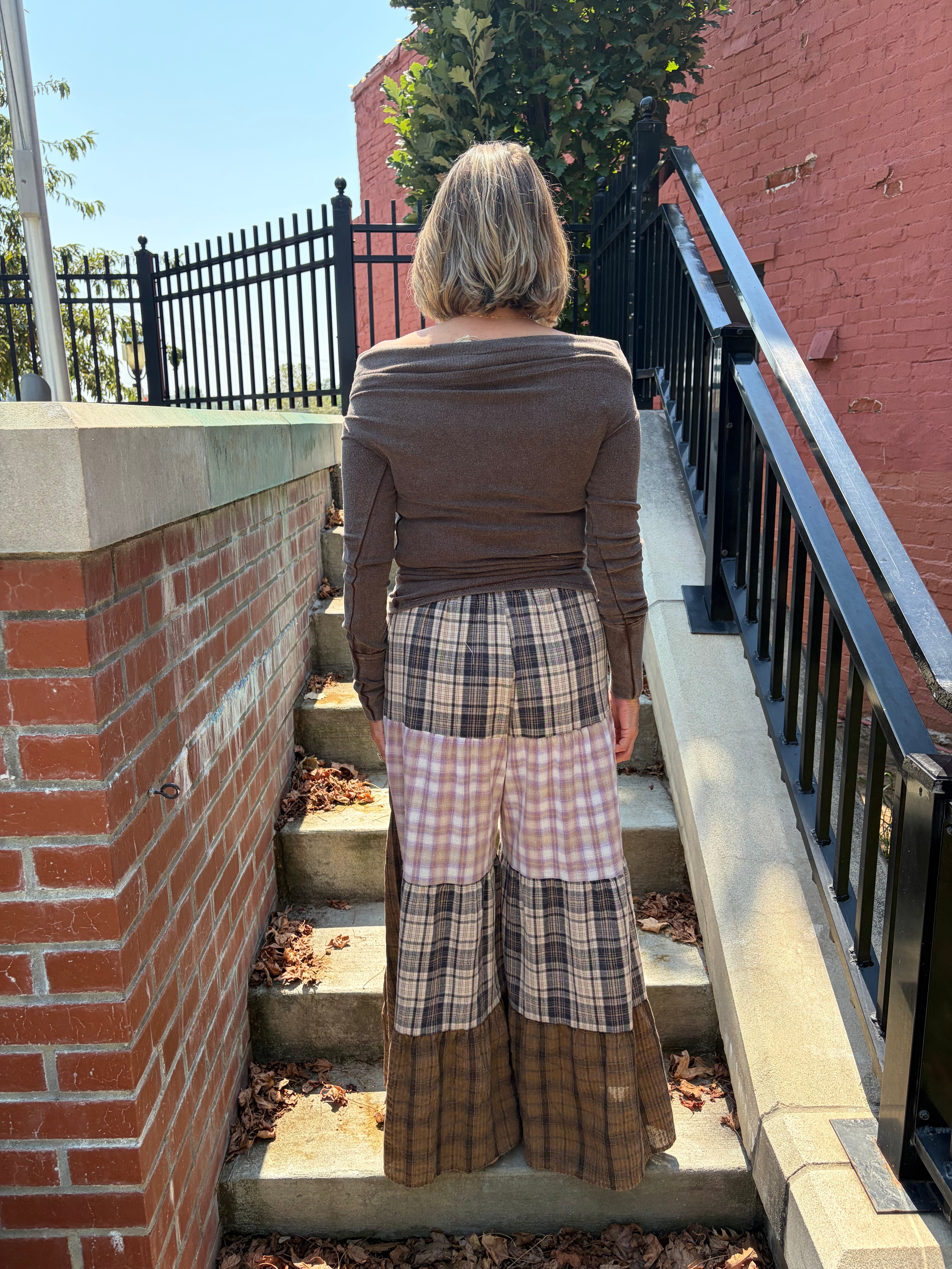 A woman viewed from behind wearing a brown off-the-shoulder long-sleeve top and a patchwork wide-leg skirt with plaid and checkered panels, standing on outdoor concrete stairs between brick walls.
