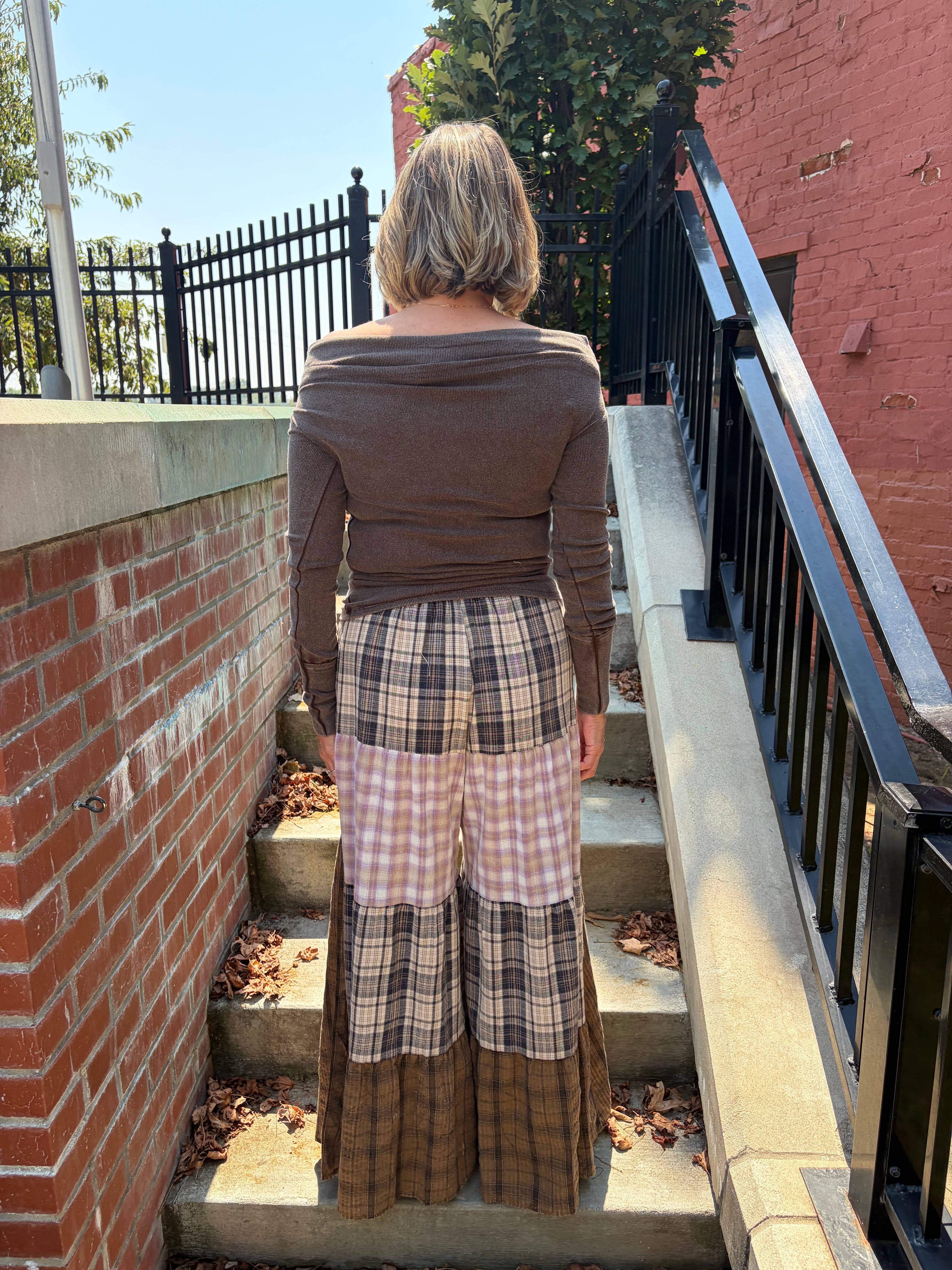 A woman viewed from behind wearing a brown off-the-shoulder long-sleeve top and a patchwork patchwork maxi skirt with plaid and gingham patterns, standing on outdoor concrete stairs between brick walls.
