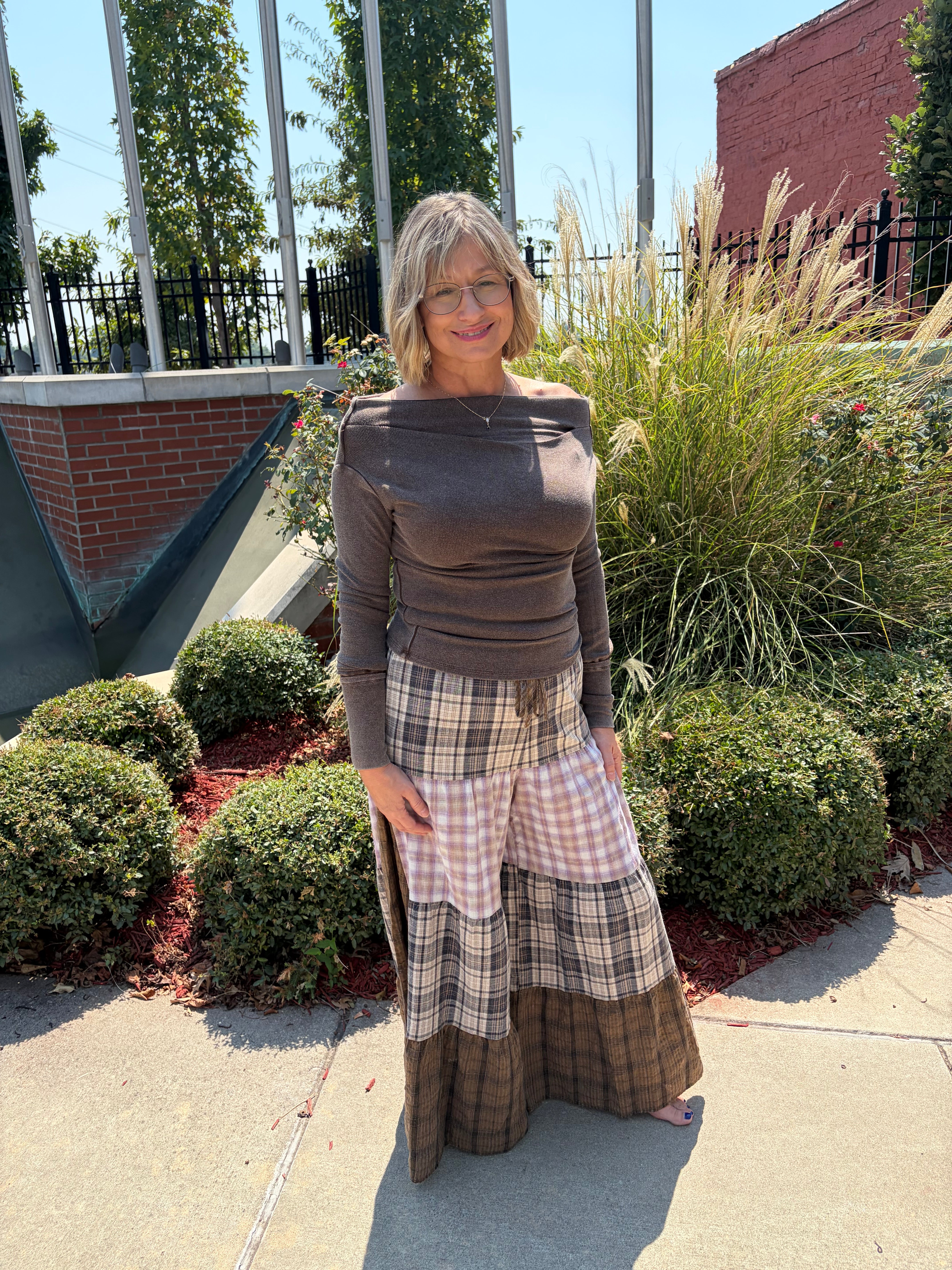 Patchwork Plaid Maxi Pants in tiered brown and neutral plaid panels, back view walking on sidewalk, styled with dark brown off-shoulder long sleeve top
