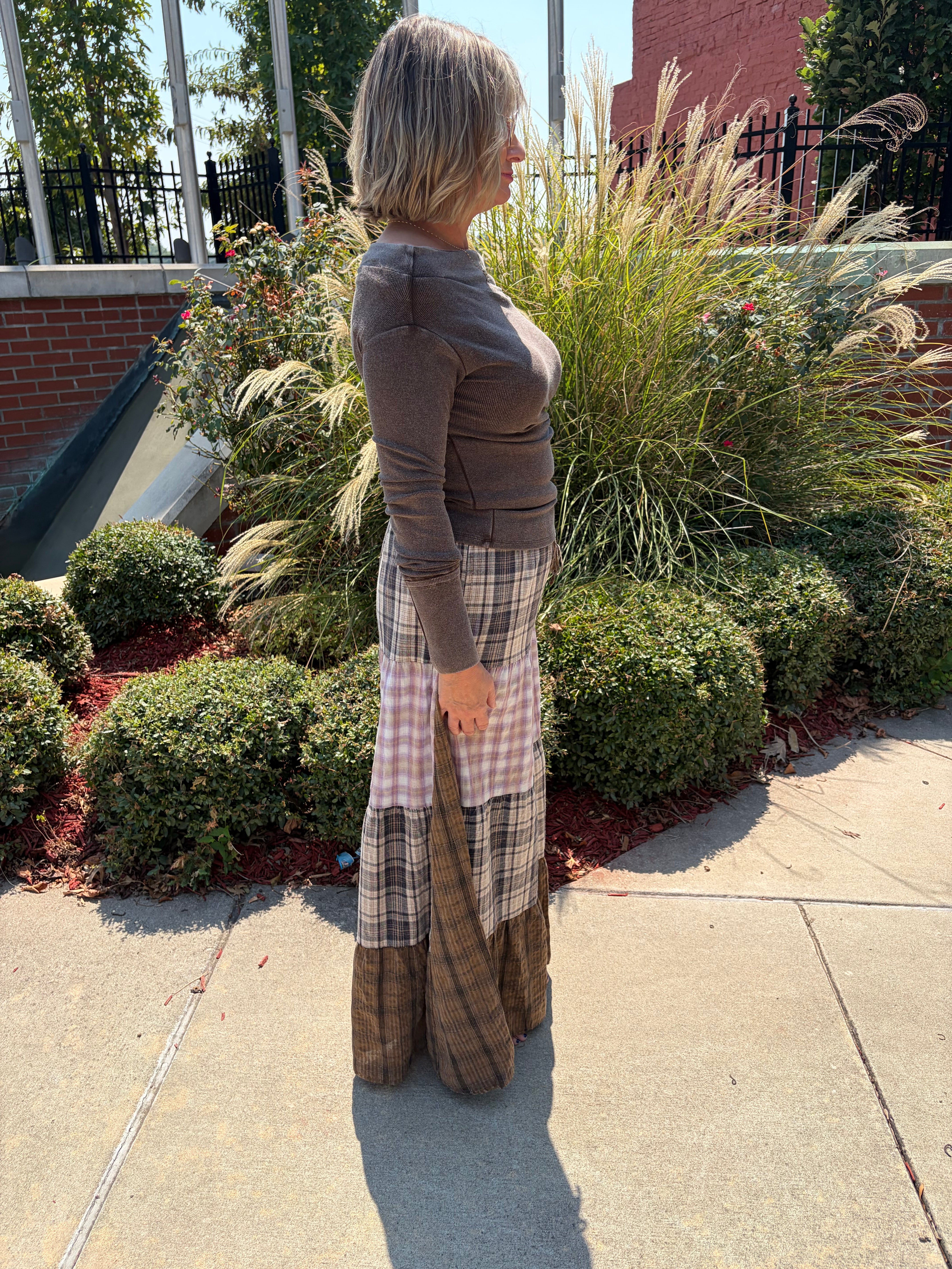 A woman stands outdoors on a sidewalk in front of landscaped bushes and ornamental grass, wearing a brown long-sleeve top and a long patchwork skirt with plaid and gingham panels.