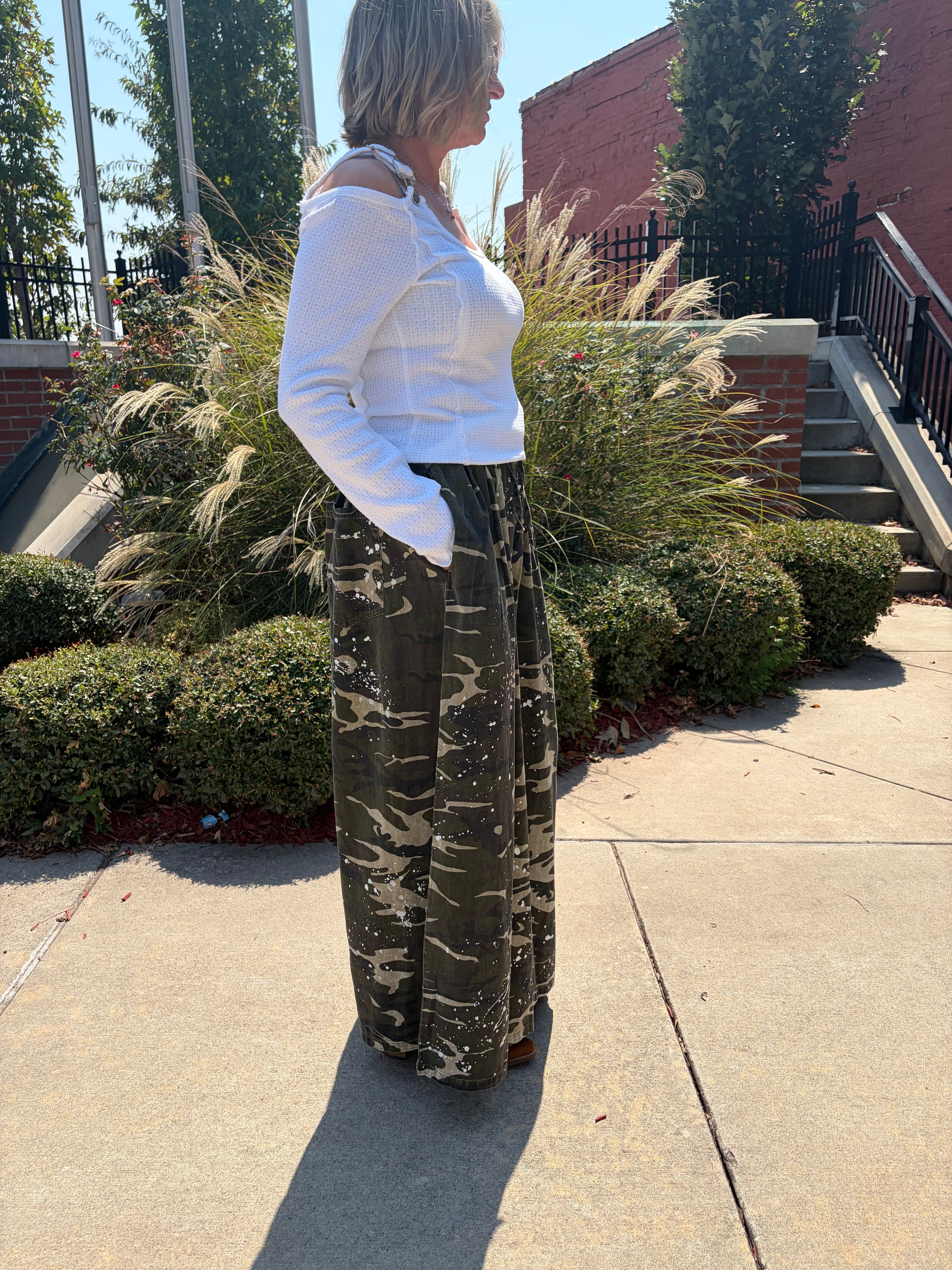 A woman standing outdoors on a sidewalk wearing a white long-sleeve top and dark olive green wide-leg pants with a camouflage pattern.