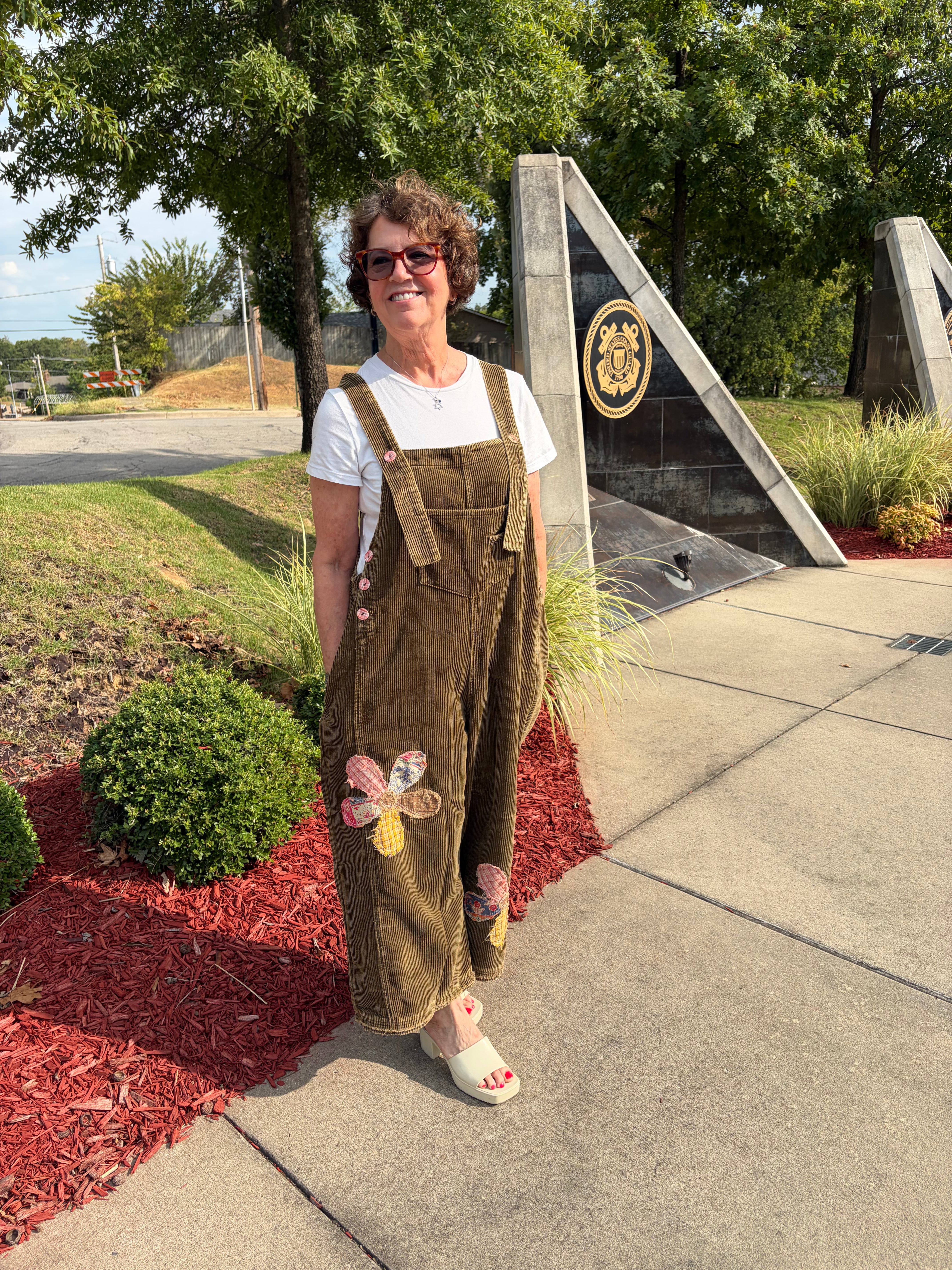 Three-quarter front view of woman wearing olive green Flower Patch Corduroy Wide-Leg Overalls with white tee and white sandals, showing floral patchwork appliqué on leg, outdoor street setting