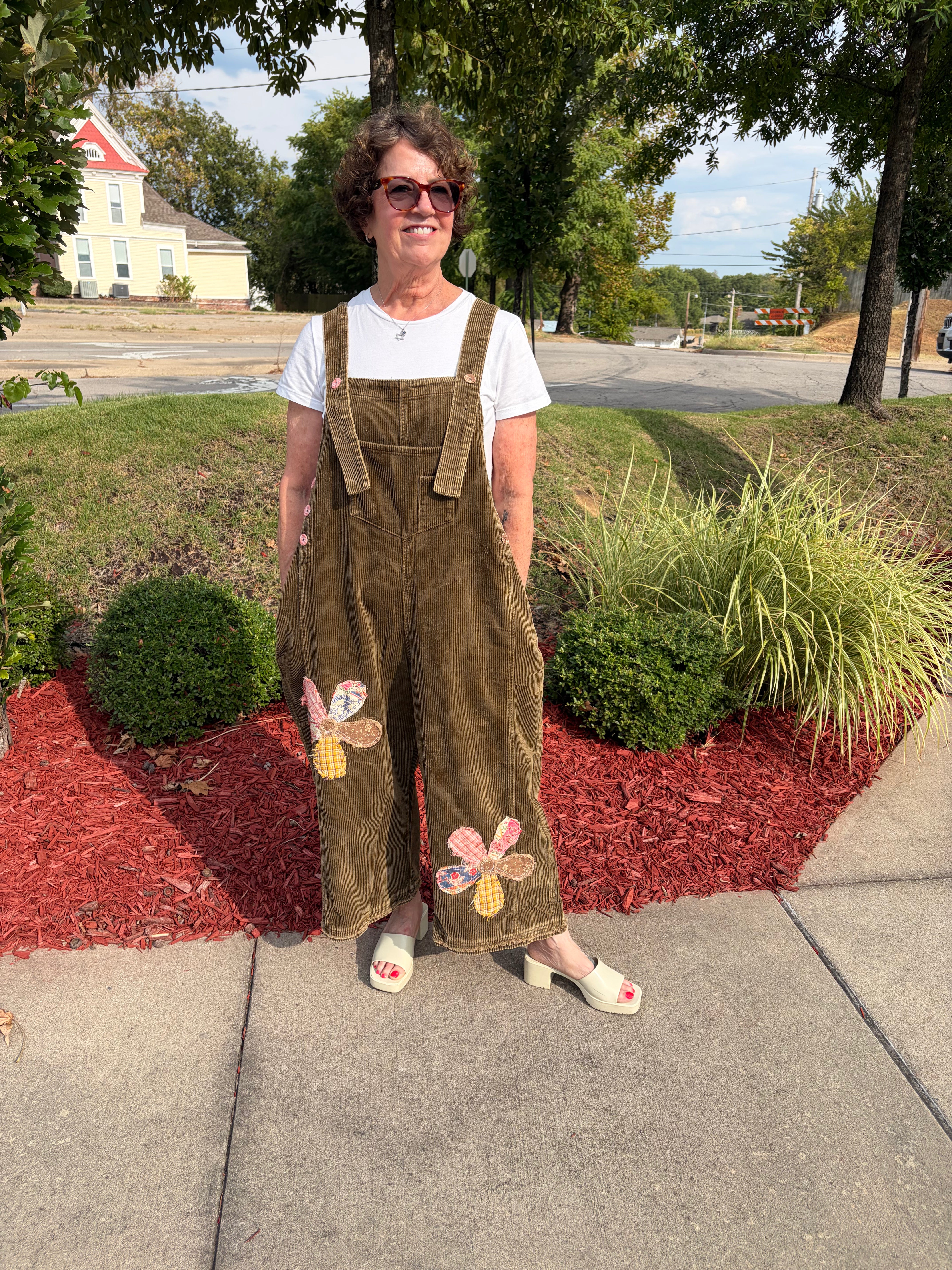 Front full-length view of woman smiling wearing olive green Flower Patch Corduroy Wide-Leg Overalls with white tee and white sandals, showing floral patchwork detail on both legs