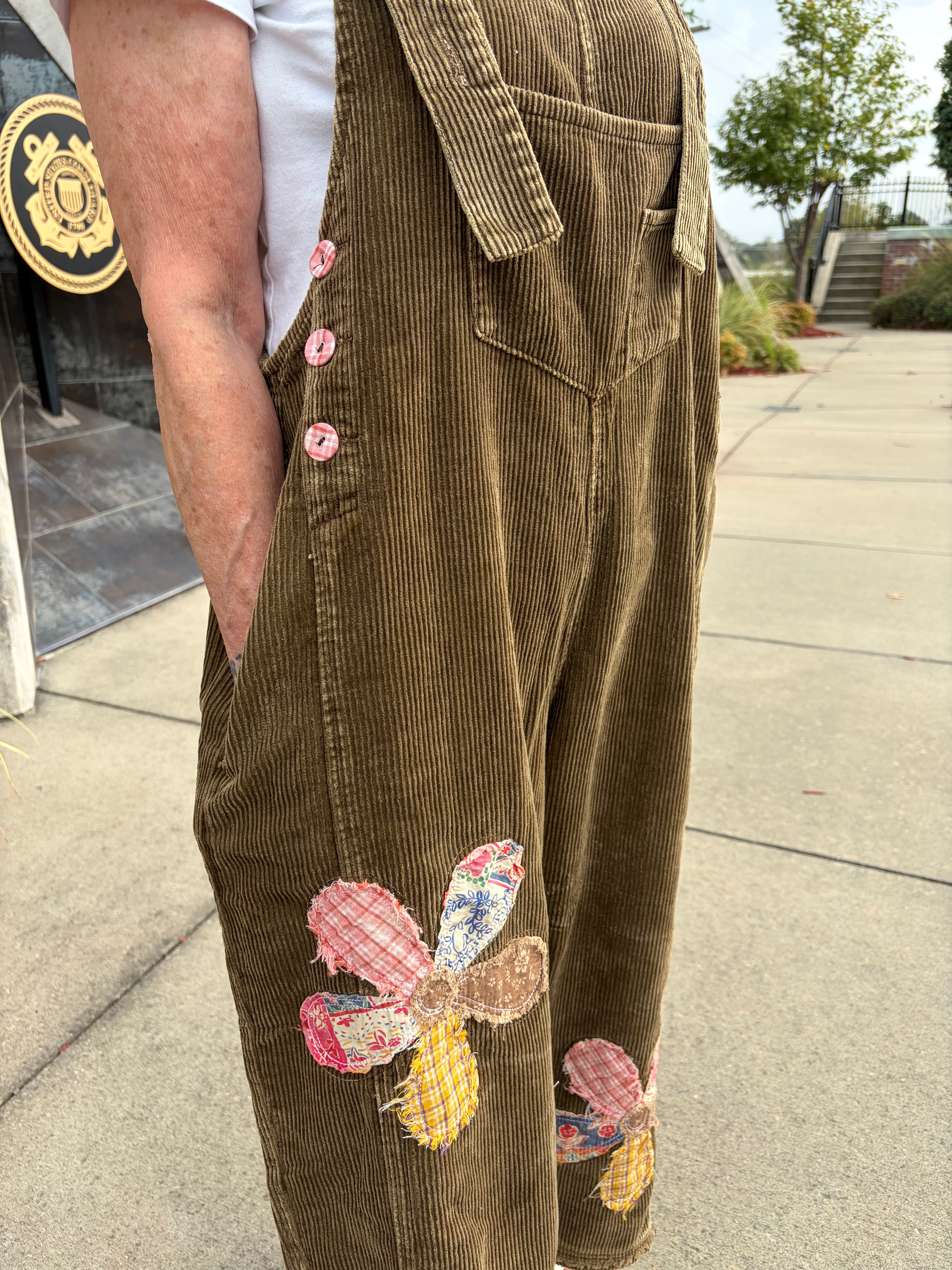 Close-up side view of olive green corduroy wide-leg overalls showing mixed-print floral patchwork appliqué on leg, side button closure, and front pocket detail