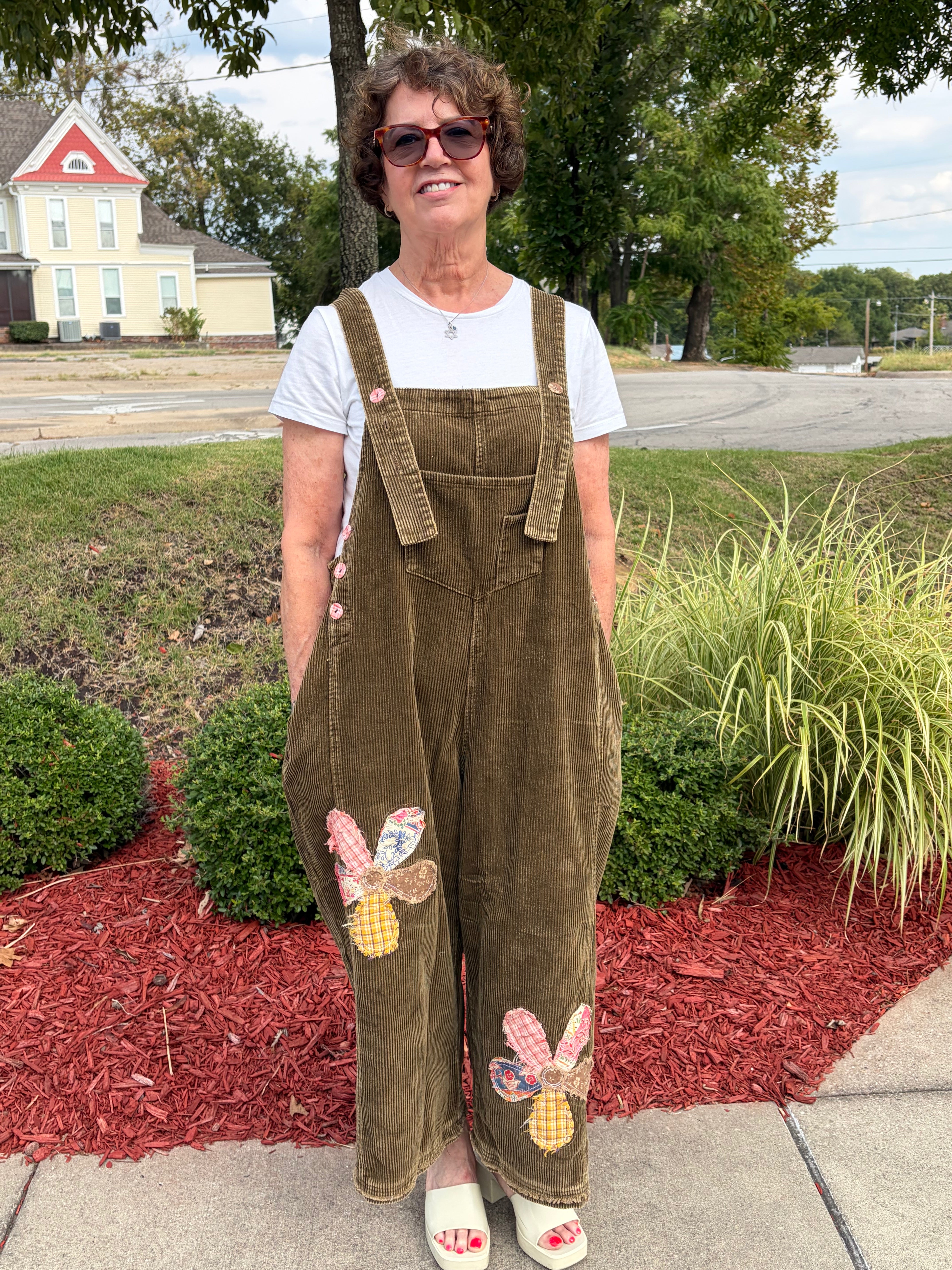 Front full-length view of woman smiling wearing olive green Flower Patch Corduroy Wide-Leg Overalls with white tee and white platform sandals, outdoor setting
