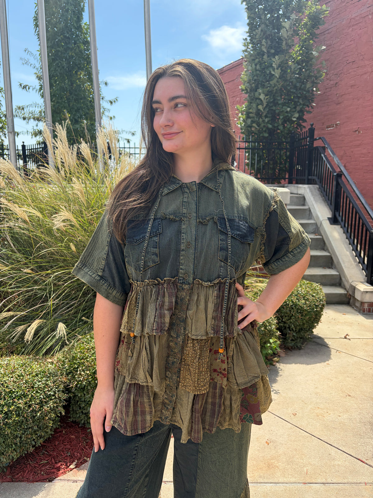A woman wearing a short-sleeve patchwork top made from olive green denim and layered ruffled brown plaid fabrics, standing outdoors on a sunny day next to a brick building and green plants.