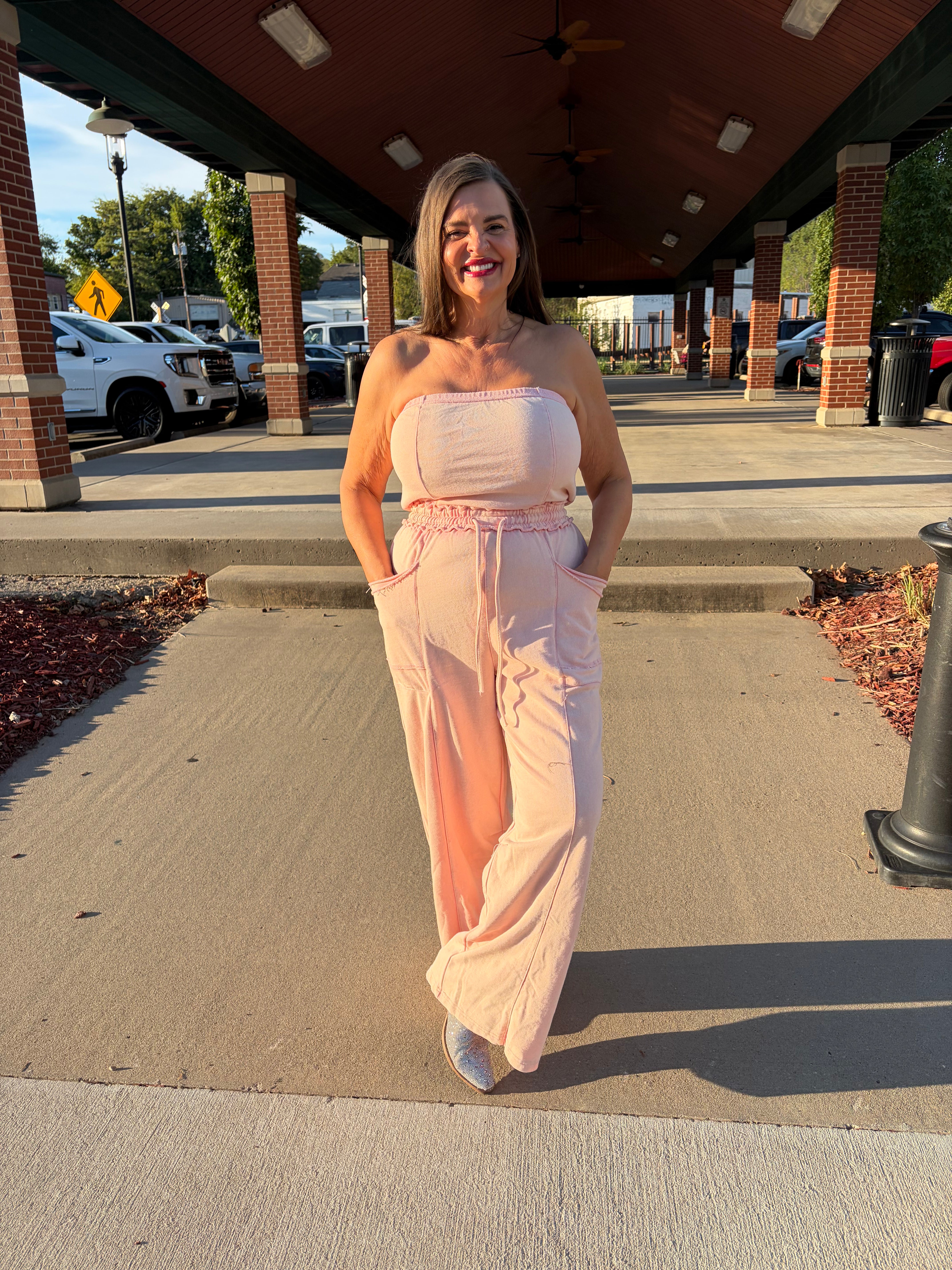 A woman wearing a light pink strapless jumpsuit walks toward the camera on a paved path under a covered outdoor pavilion.