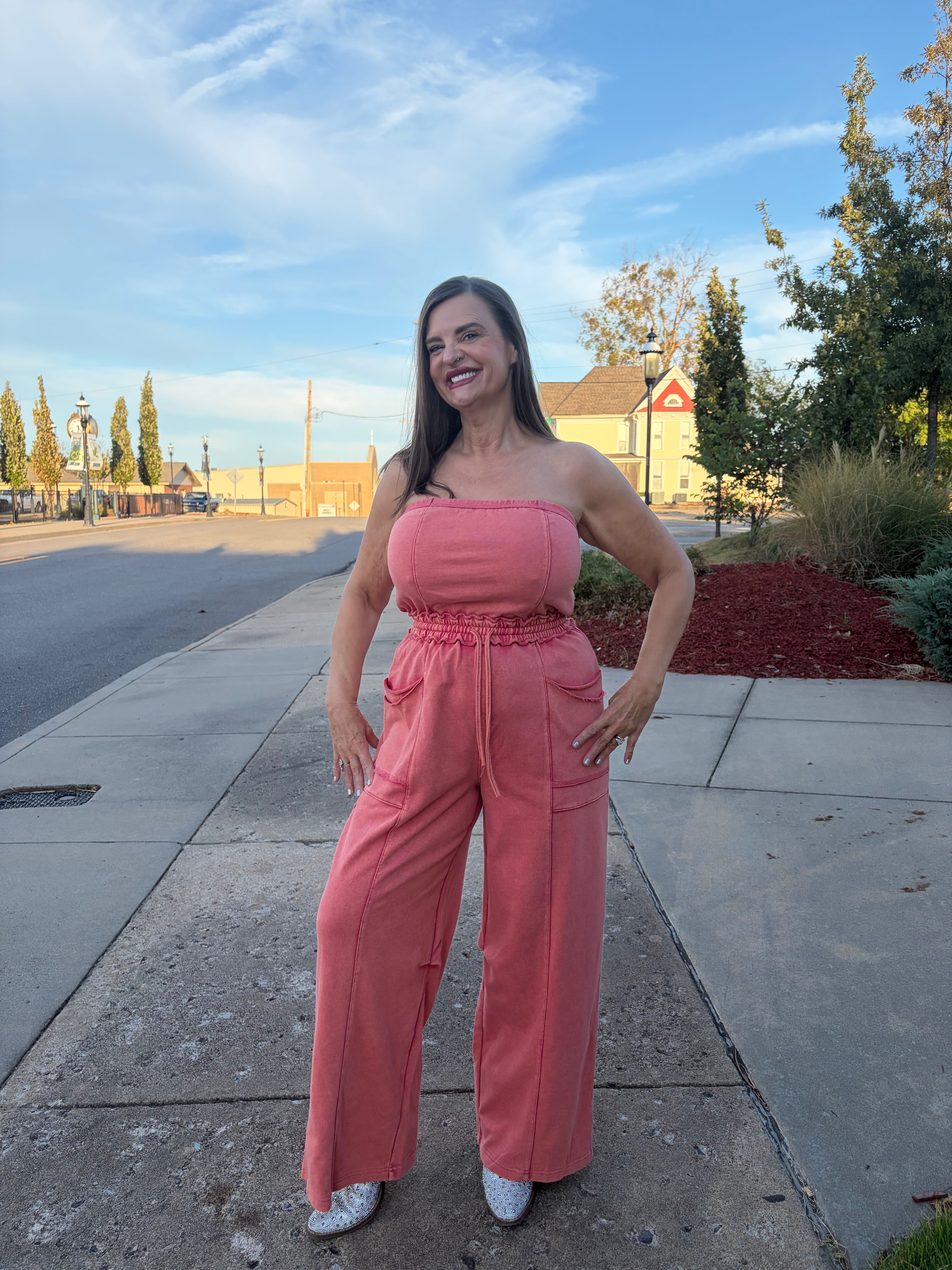 A woman modeling a pink strapless jumpsuit with wide legs and a tie waist, standing on a concrete sidewalk outdoors.