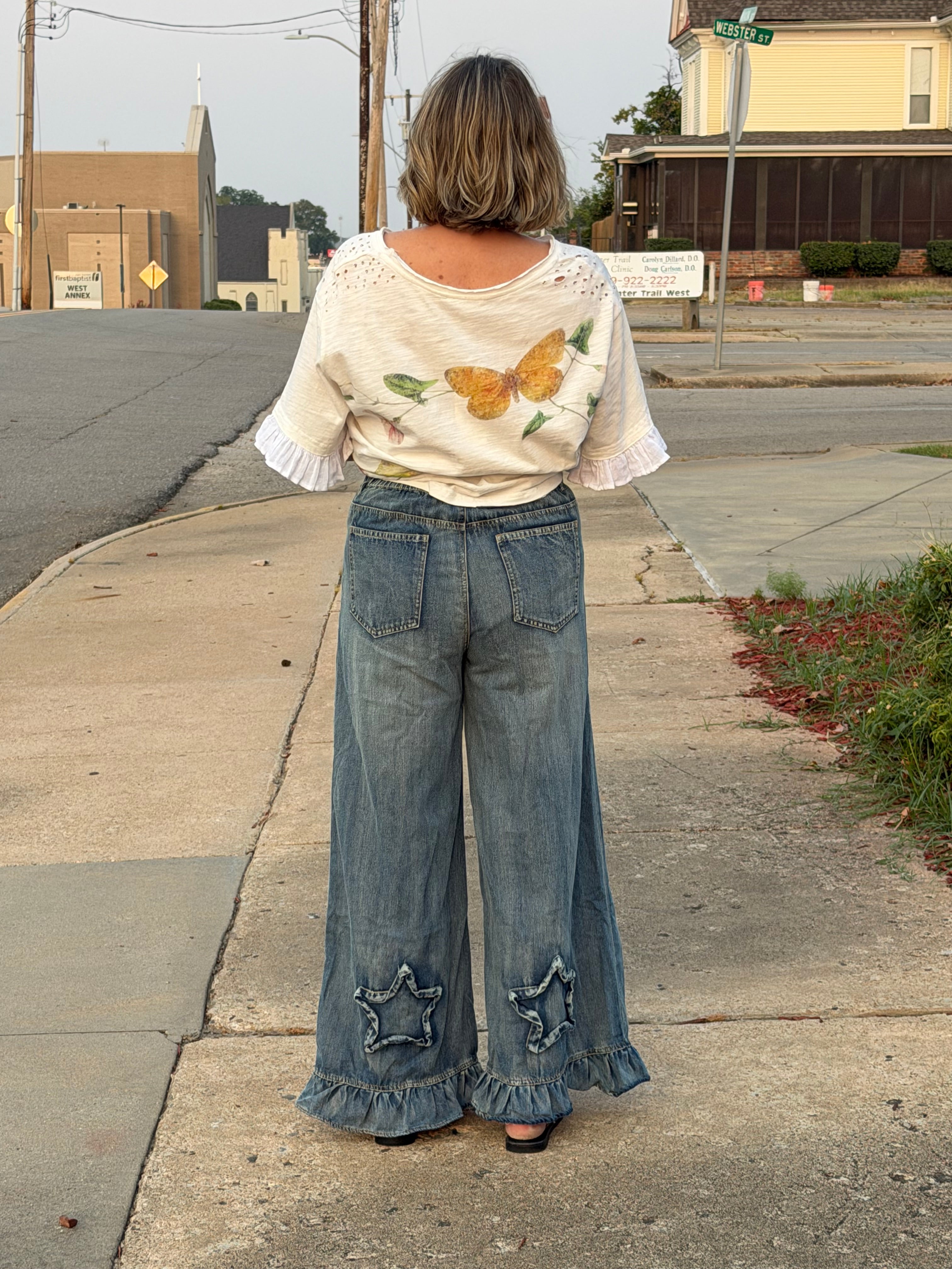 A woman stands on a sidewalk outdoors, viewed from behind, wearing a cream short-sleeve top with butterfly embroidery and wide-leg blue jeans with ruffled hems and star patch details.