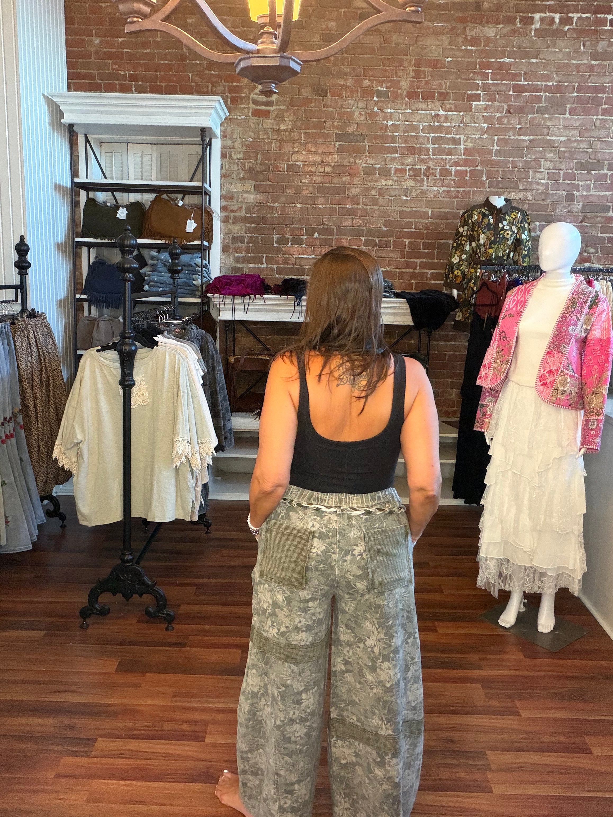 A woman stands with her back to the camera inside a boutique, wearing a black tank top and wide-leg camouflage pants. The shop has a brick wall, wooden floors, clothing racks, and mannequins displaying other outfits.