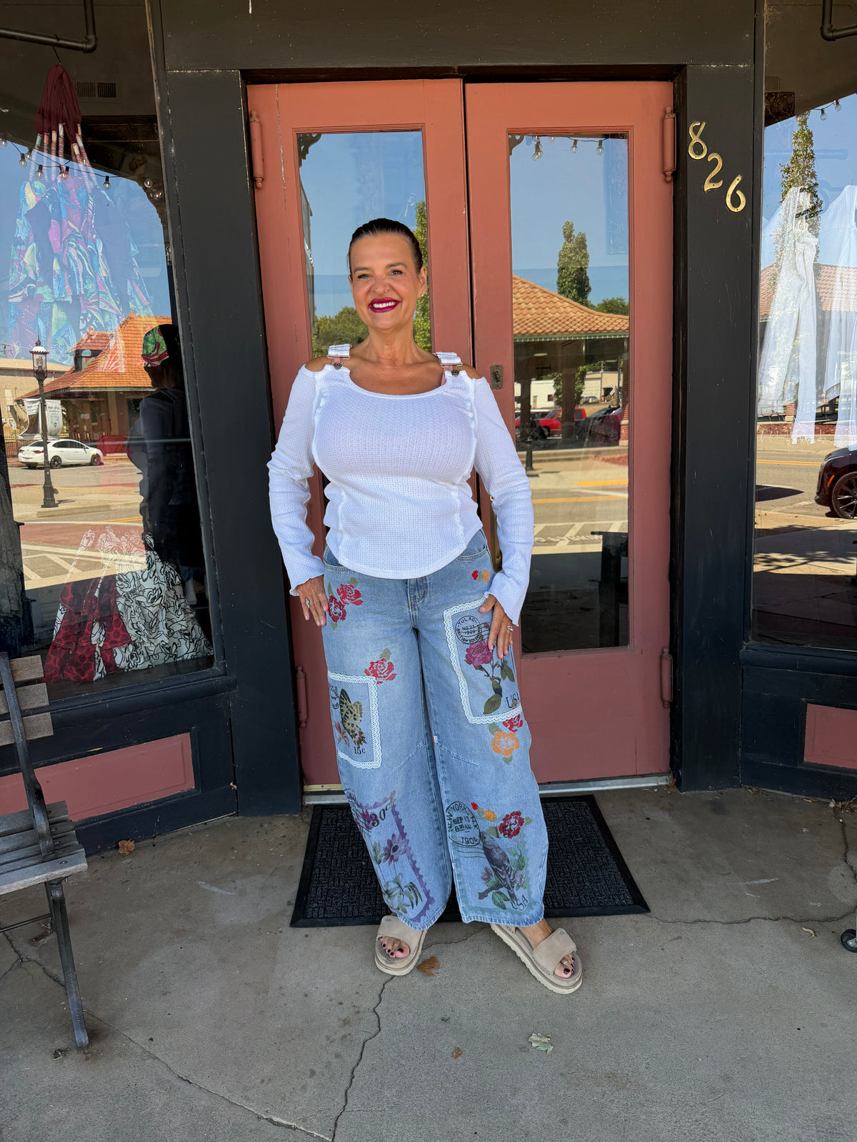 Woman smiling in Alissa Patchwork Pant in light blue denim with floral and stamp patchwork, full body front view styled with rust orange boho blouse and beige heeled booties, boutique storefront