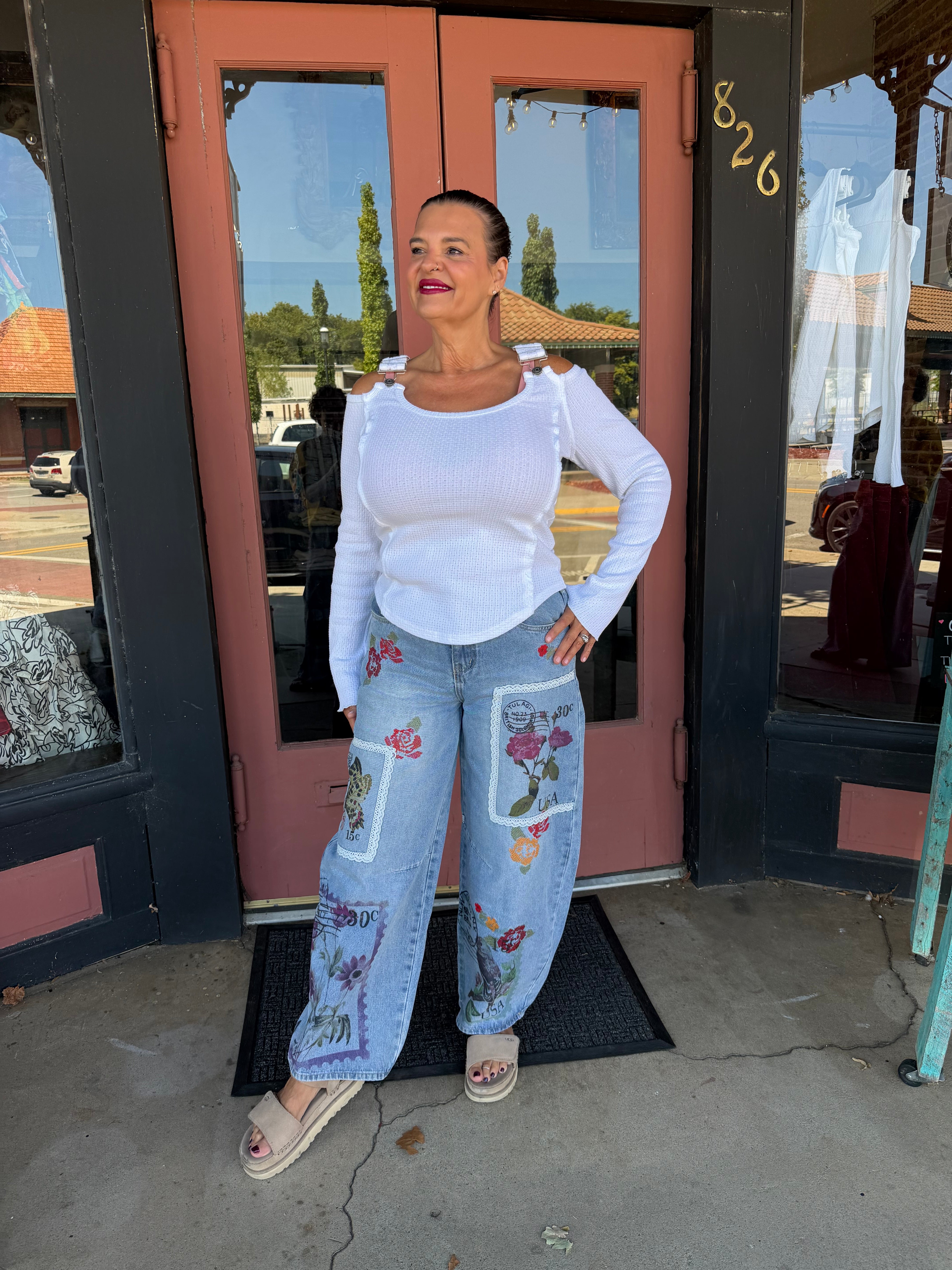 Woman smiling in Alissa Patchwork Pant in light blue denim with floral and stamp patchwork, full body front view styled with rust orange boho blouse and beige heeled booties, boutique storefron
