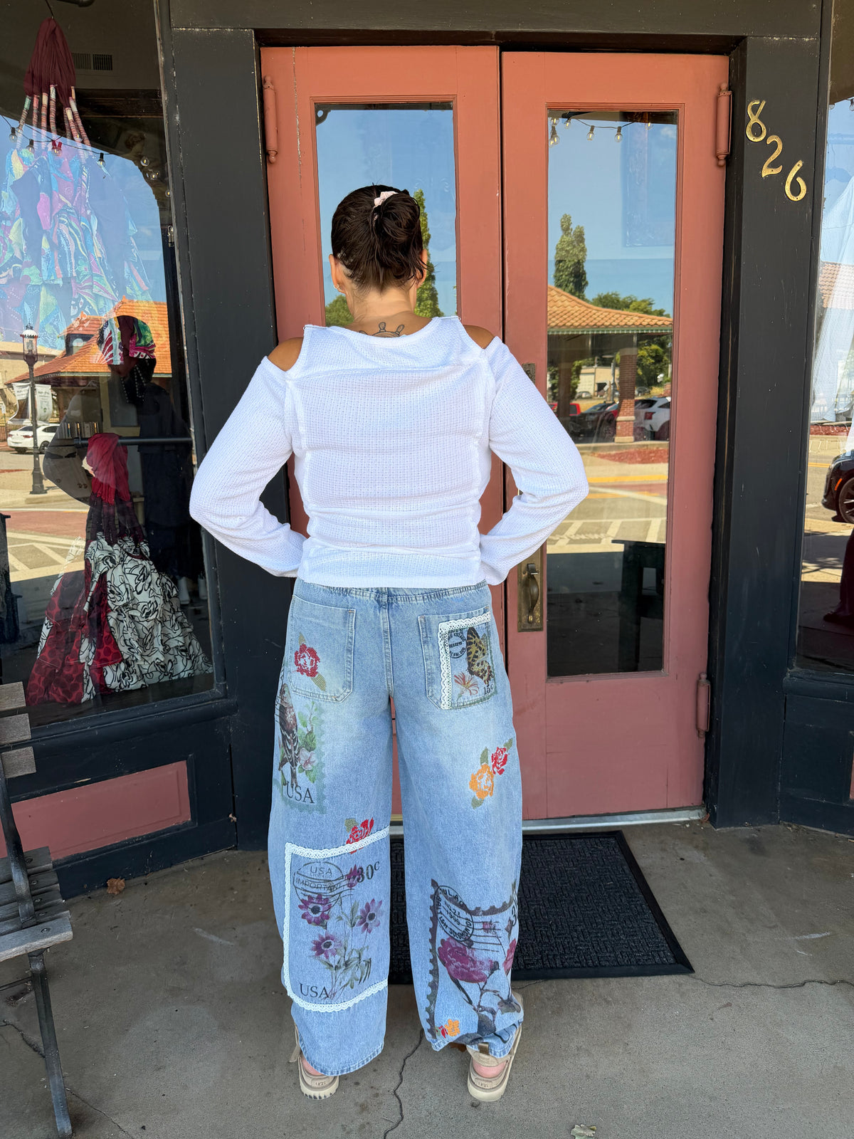 Back view Woman smiling in Alissa Patchwork Pant in light blue denim with floral and stamp patchwork, full body front view styled with rust orange boho blouse and beige heeled booties, boutique storefront