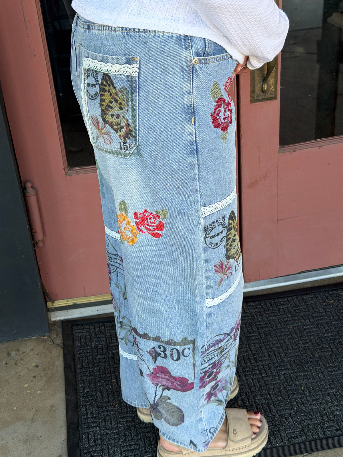 side view Close-up of Alissa Patchwork Pant back pockets and leg panels featuring colorful butterfly, rose, and vintage postage stamp embroidery on light blue denim