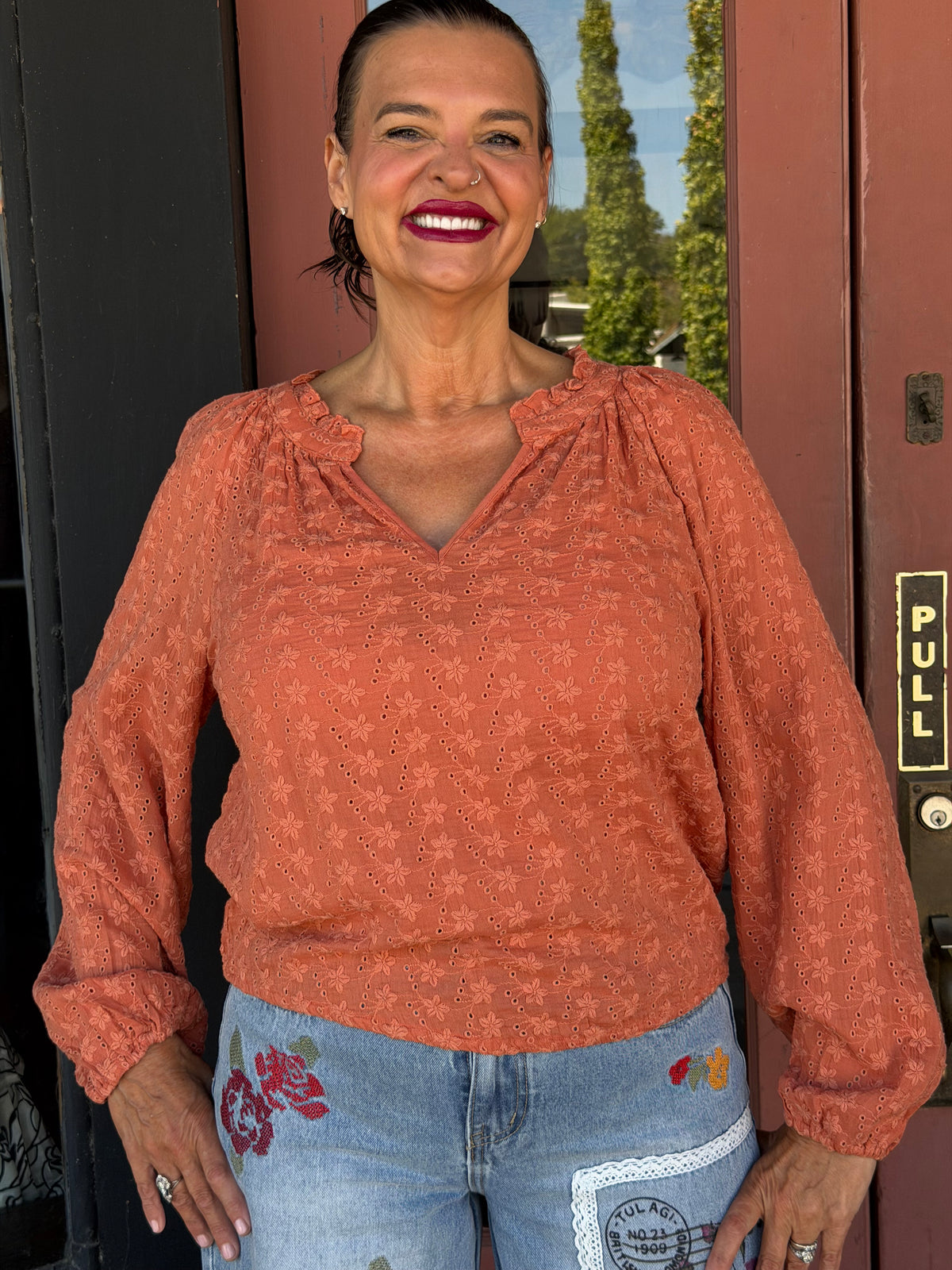 Woman smiling in a terracotta floral eyelet long sleeve blouse with ruffle V-neckline, styled with embroidered floral jeans, front view outdoors