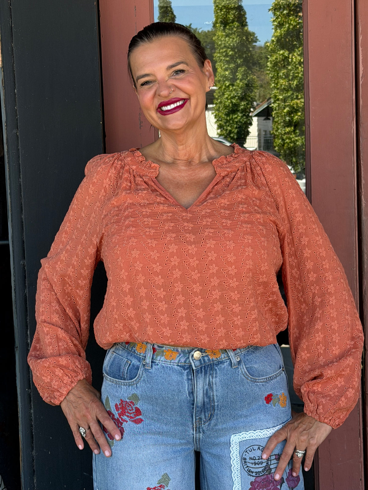 Woman smiling in a terracotta floral eyelet blouse with ruffle V-neckline and elastic cuffs, front close-up view, boutique doorway

