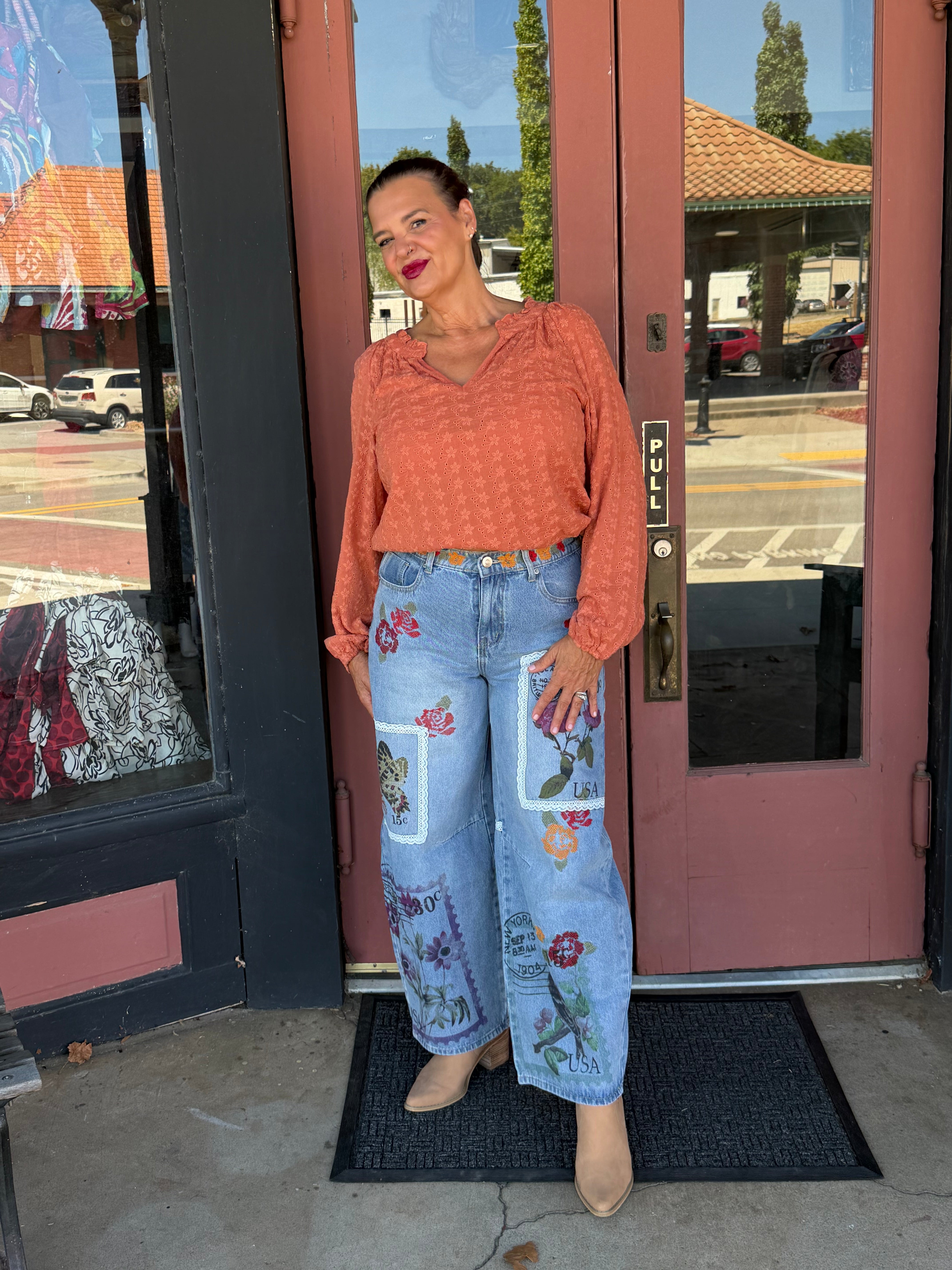 Woman smiling in Alissa Patchwork Pant in light blue wash with floral and postage stamp embroidery patches, three-quarter front view styled with white cold-shoulder ribbed top and beige platform sandals, boutique storefront