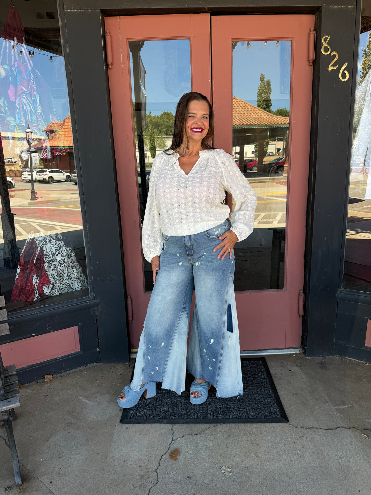 Woman smiling in Jolene Wide Leg Flare Jeans in medium blue wash with paint splatter distressing and patchwork godet panels, full body front view, boutique exterior