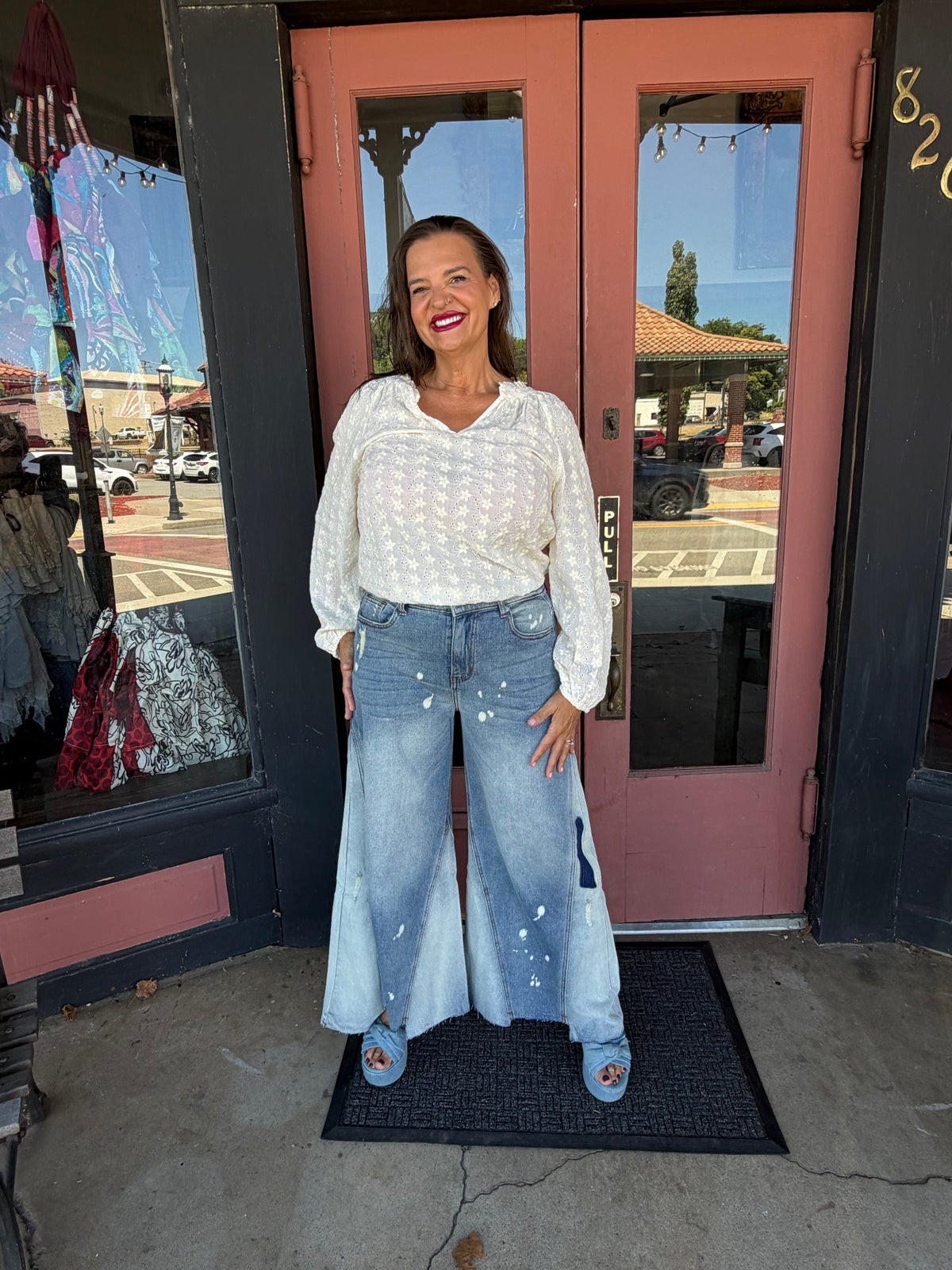 Woman smiling in Jolene Wide Leg Flare Jeans with paint splatter detailing and wide godet flare, full body front view styled with white eyelet blouse and blue platform heels, boutique doorway
