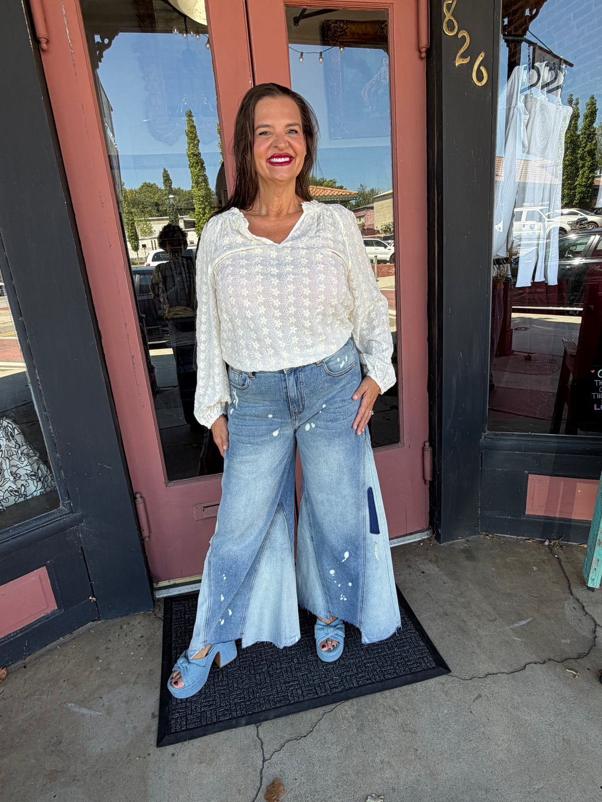 Woman smiling in a white floral eyelet blouse with ruffle neckline, full body front view styled with wide-leg jeans and blue heels, boutique exterior