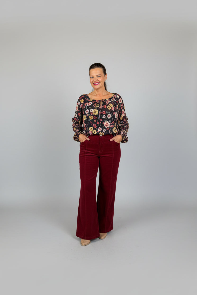 A woman stands in front of a storefront doorway wearing a floral long-sleeve top and burgundy wide-leg pants. The photo is taken outdoors on a sidewalk with the pink double doors and street reflections visible.