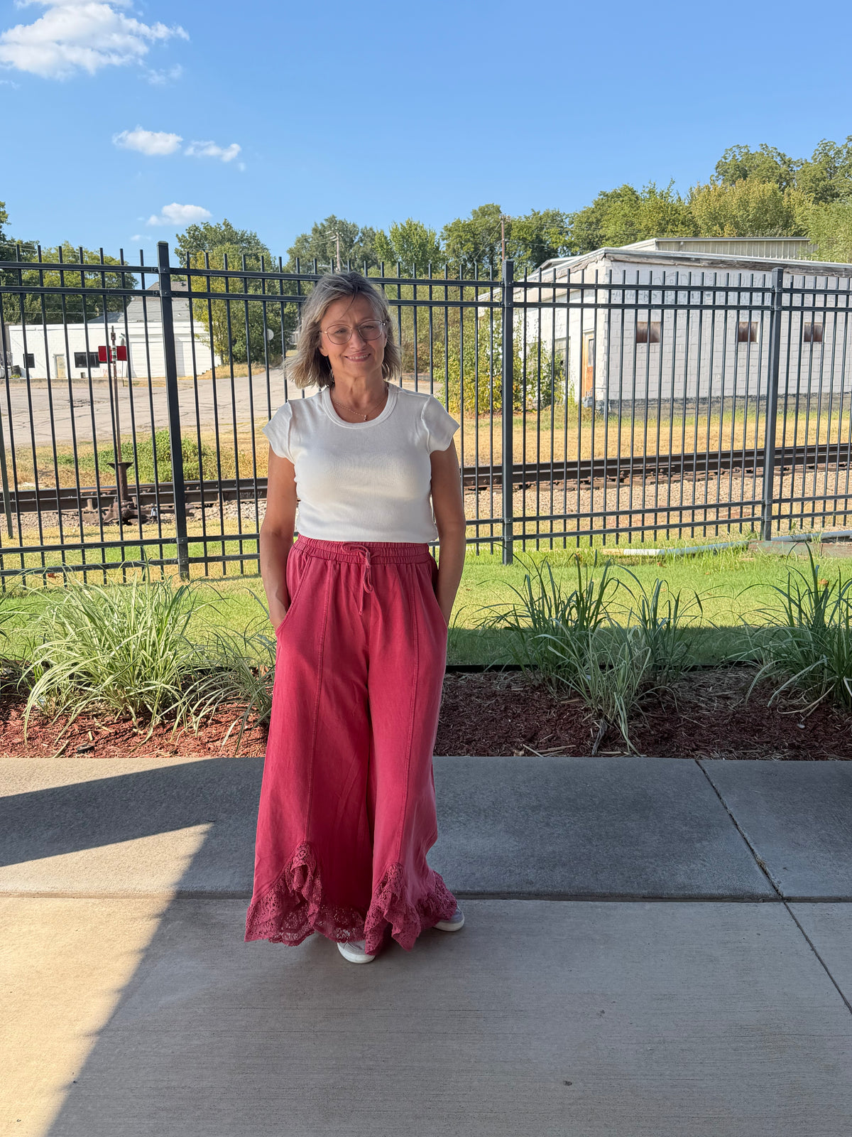 A woman stands outdoors on a concrete patio wearing a white short-sleeve crewneck t-shirt and high-waisted, wide-leg dusty rose pink pants with a ruffled hem. Behind her is a black metal fence, green grass, and trees under a blue sky.