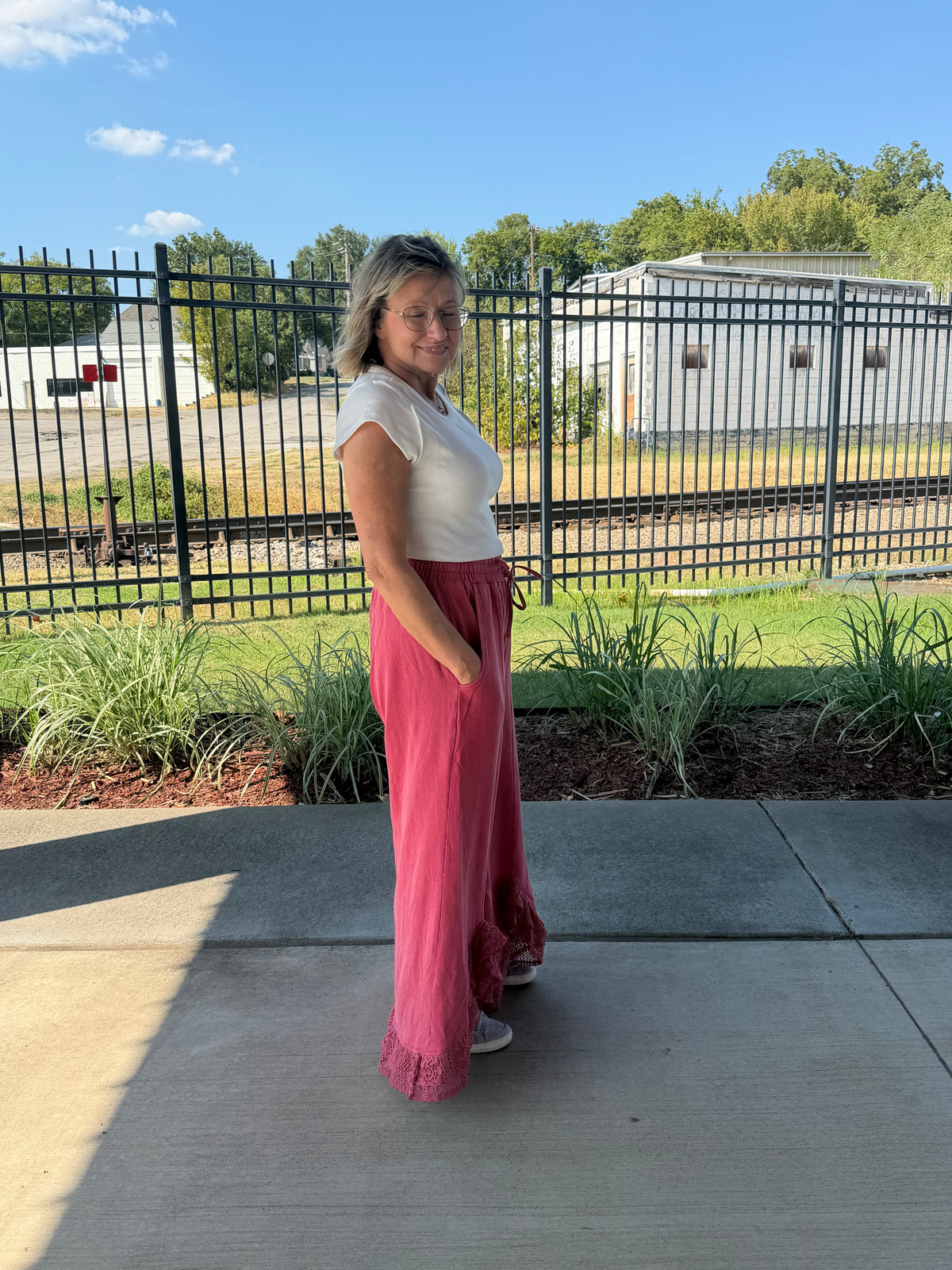 A woman stands outdoors on a concrete patio wearing a white short-sleeve top and a long, flowing dusty pink skirt or pants. Behind her is a black metal fence, green grass, trees, and a blue sky.