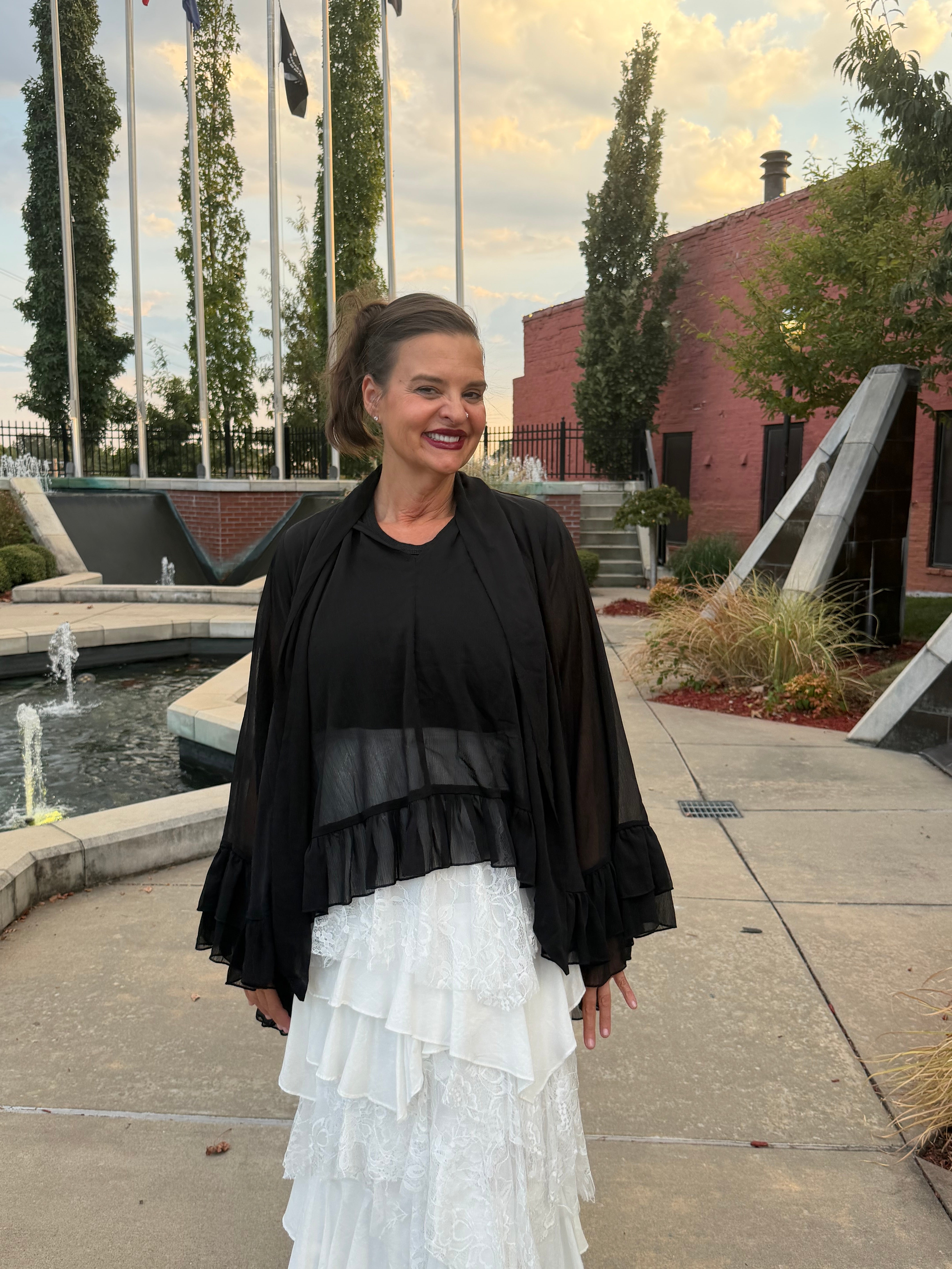 A woman stands outdoors wearing a black sheer long-sleeve top layered over a white tiered ruffled skirt. The background shows a paved walkway, a fountain, flagpoles, trees, and a brick building under a cloudy sky.