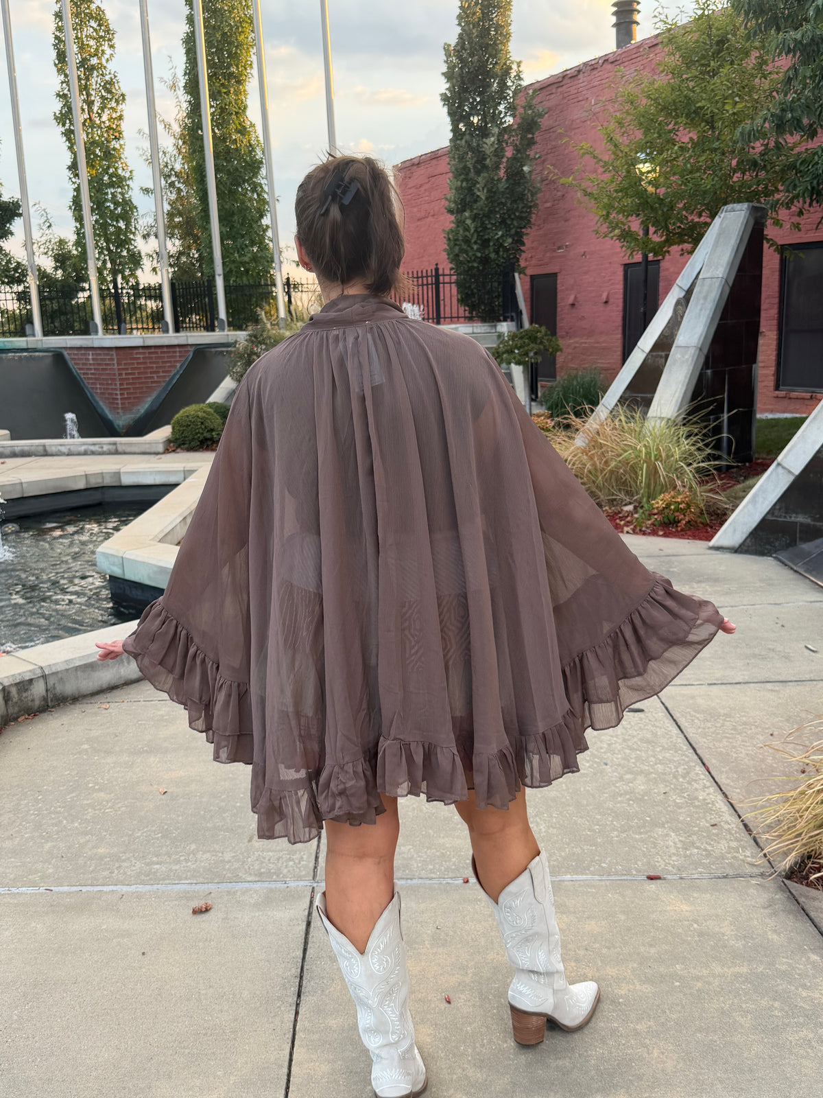 A woman viewed from behind wearing a sheer taupe-brown ruffled cape-style dress or poncho outdoors on a concrete walkway, with white cowboy boots and a brick building in the background.